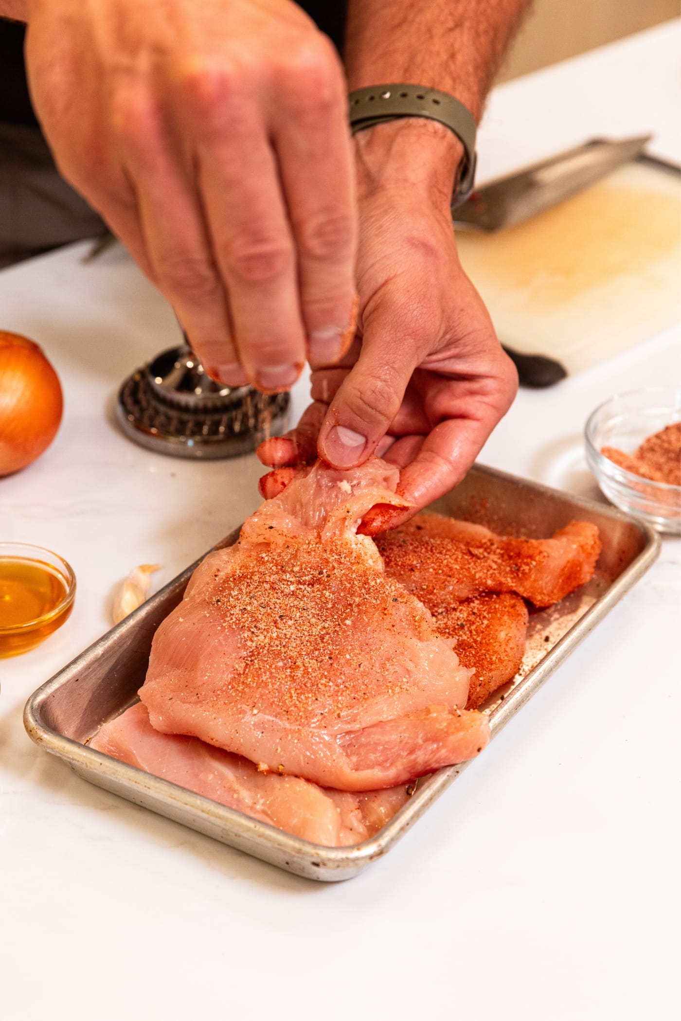 Hand holding onto piece of raw chicken sprinkling with dry spice rub on small metal baking pan.