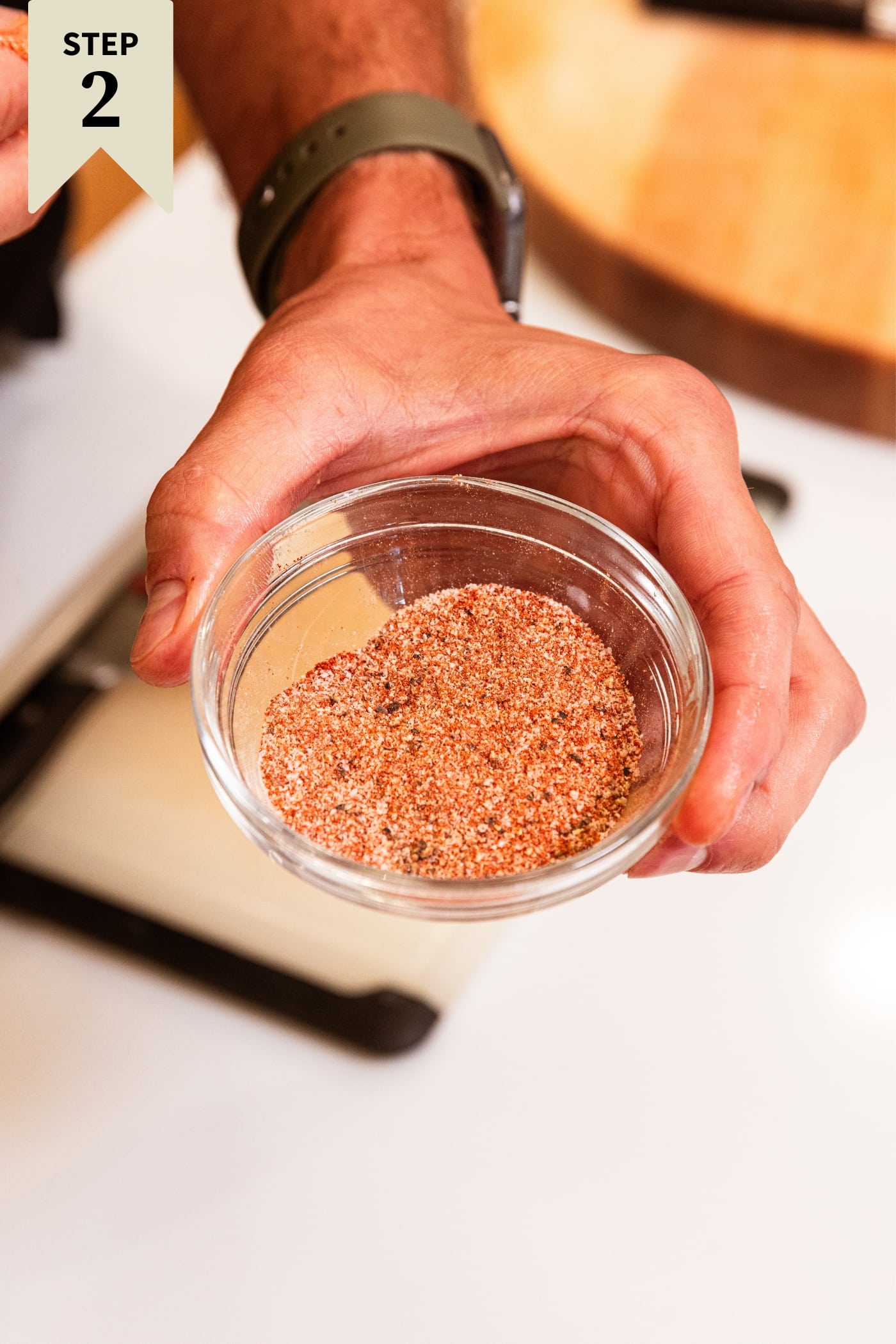 Hand holding small glass bowl filled with mixture of spices brown in color with cutting board below.