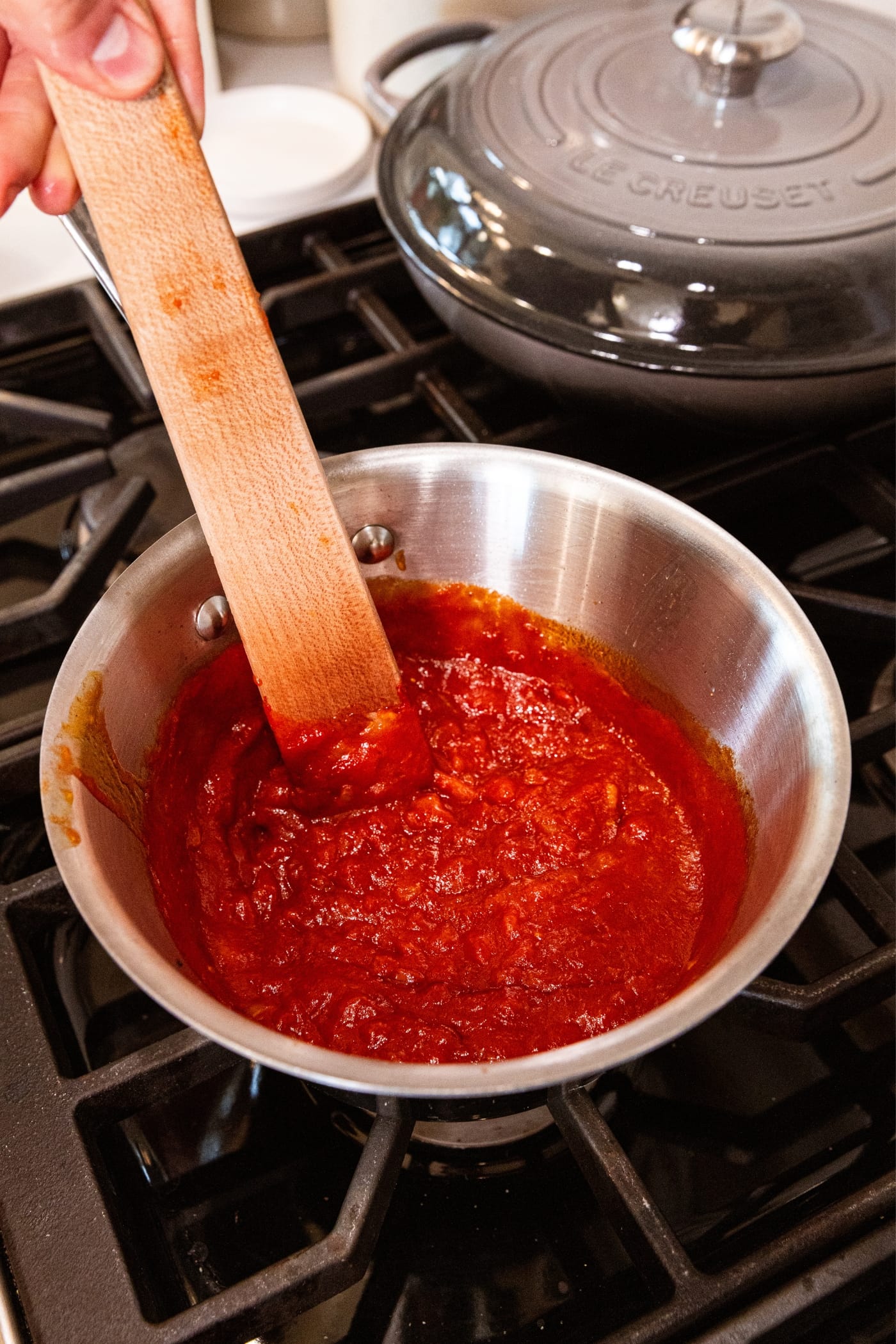 Wooden spatula stirring together red colored barbecue sauce in small saucepan sitting on stovetop.
