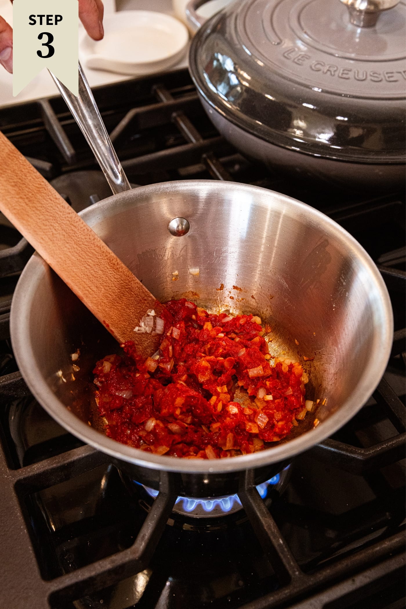 Saucepan sitting on stovetop with cooking mixture of tomato paste and diced onion with wooden spatula mixing everything together.