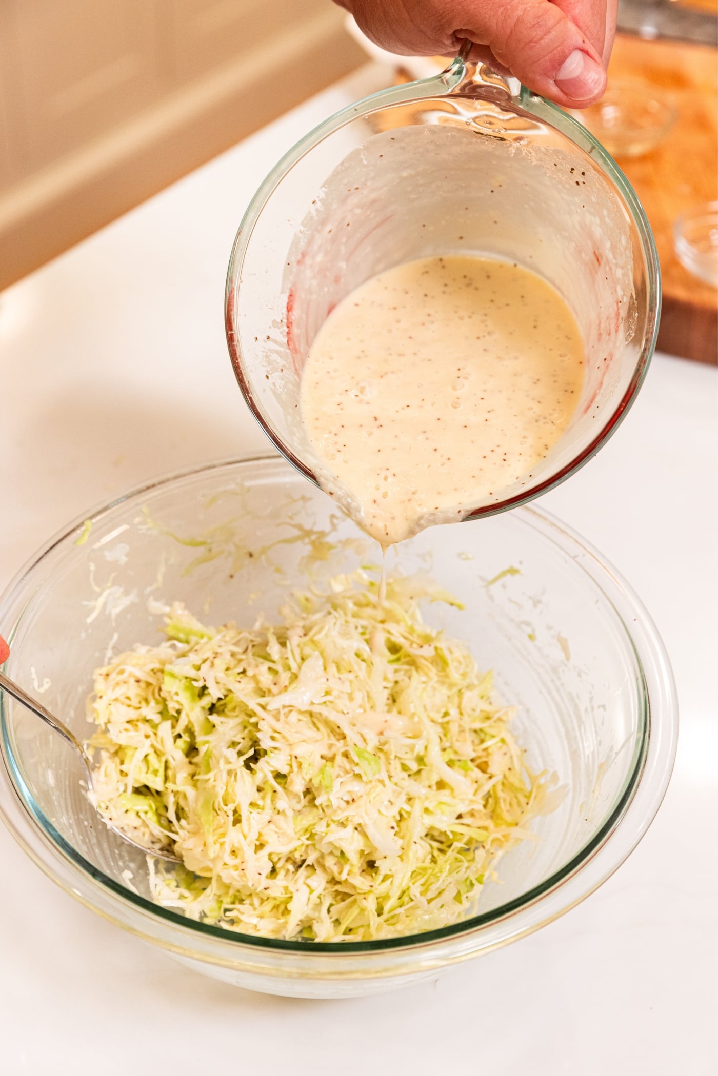 Hand holding glass measuring cup filled with white colored onion dressing pouring on top of shredded cabbage in a bowl.