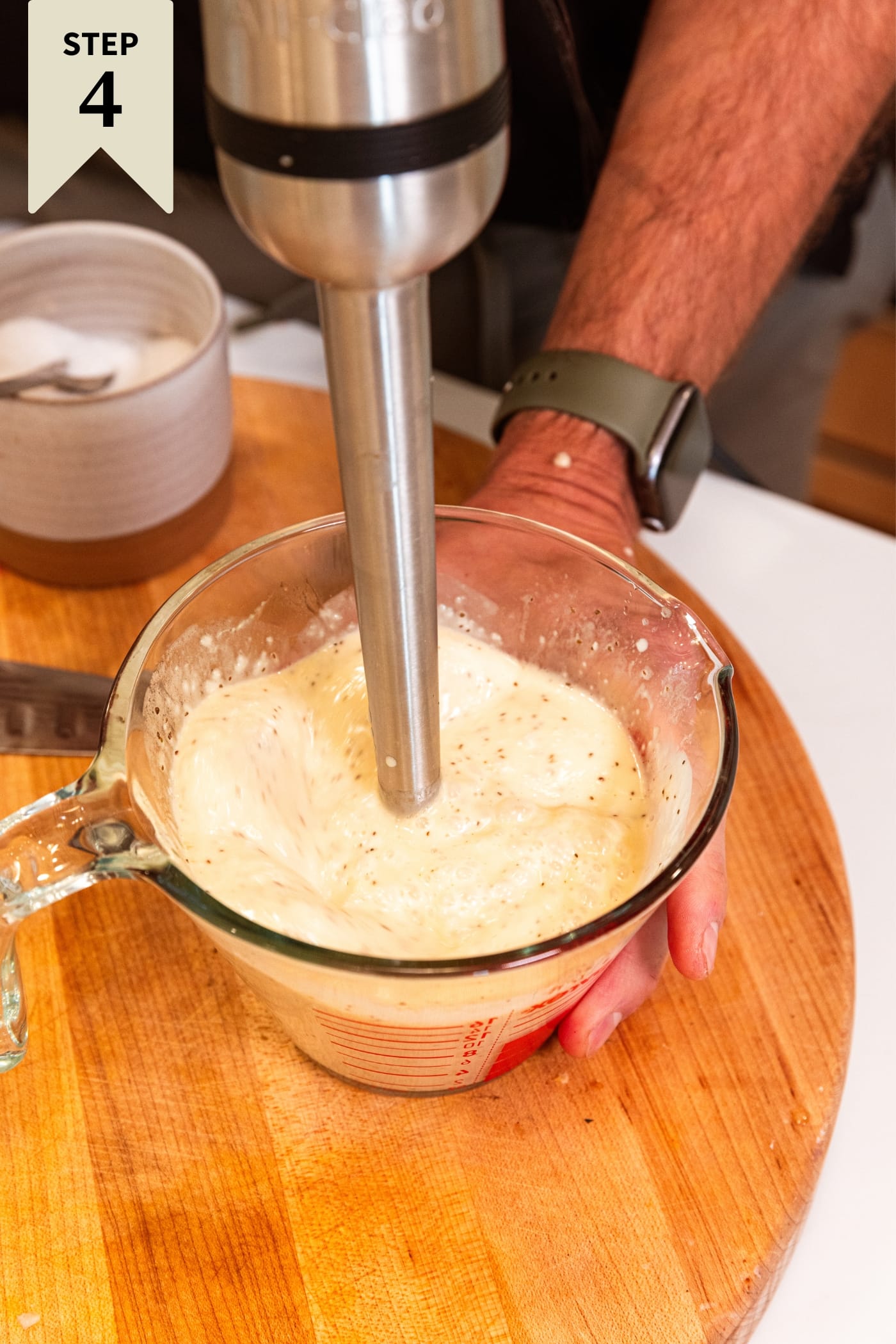 Hand holding immersion blender blending together white onion and celery seed mixture in glass measuring cup on cutting board.
