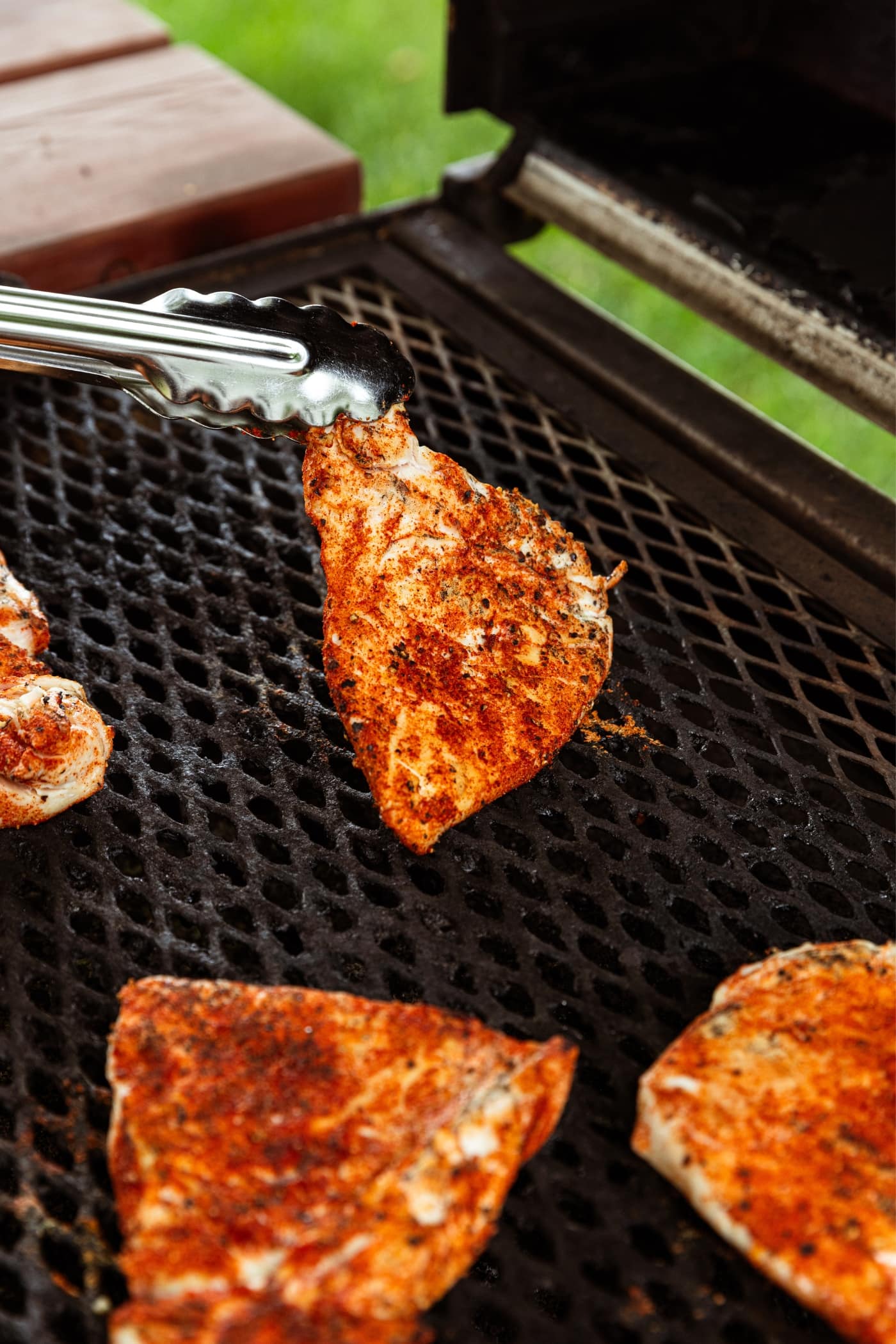 Hand holding tongs turning over grilling piece of chicken breast on grill grates.