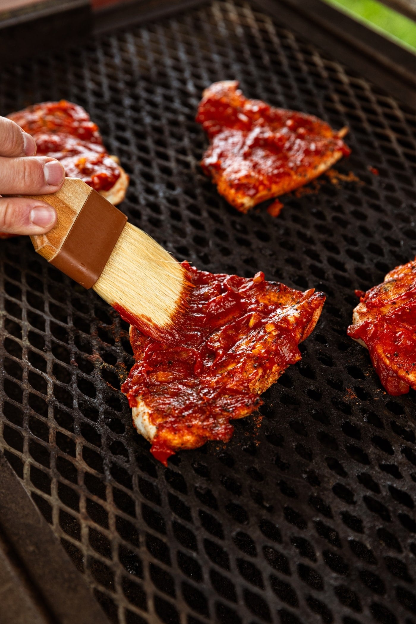 Hand holding small brush brushing barbecue sauce onto chicken breasts on the surface of a grill.