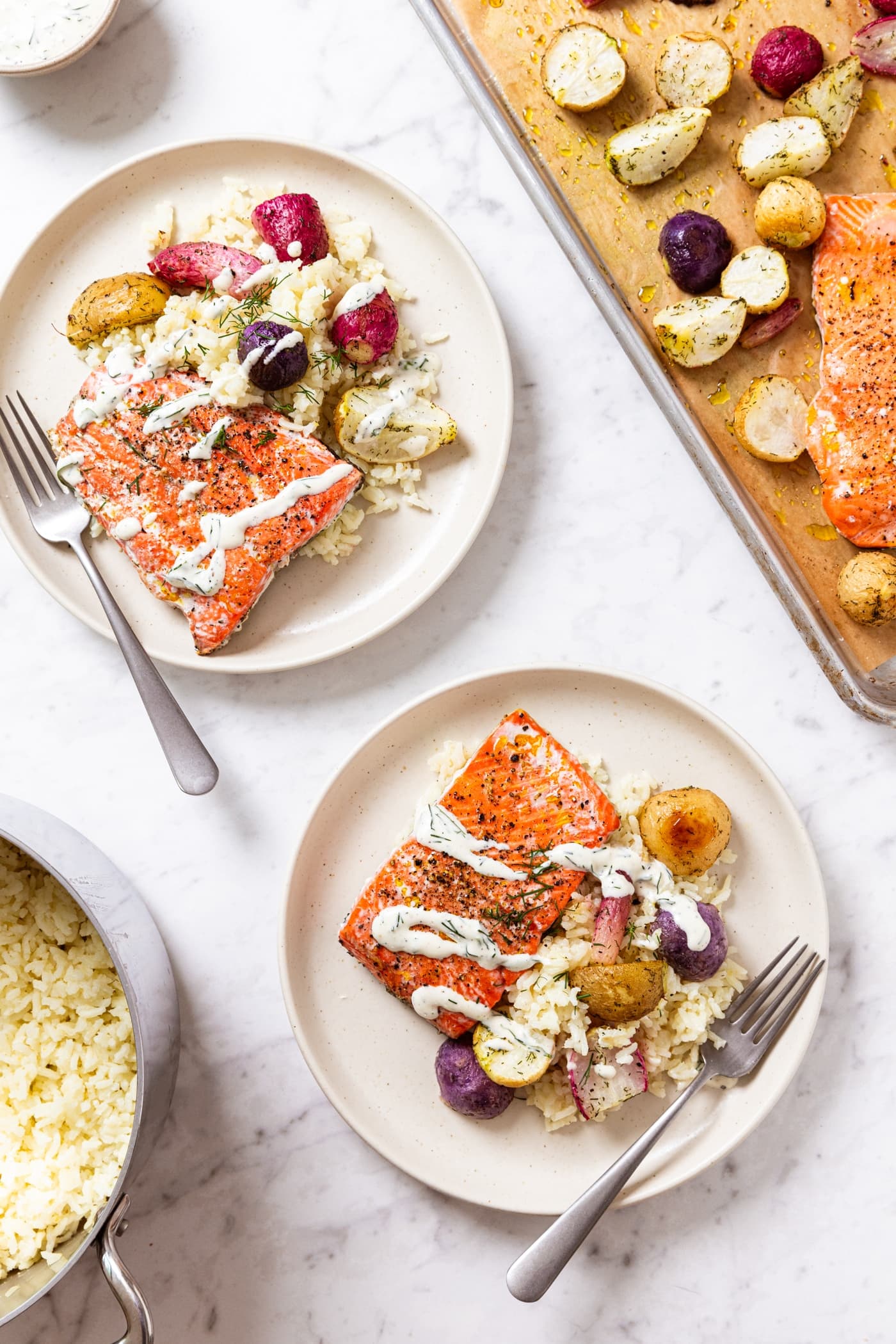White marble surface with two plates filled with salmon filets, sliced radishes, cooked rice, and drizzled with ranch dressing with baking sheet with rest of radishes.