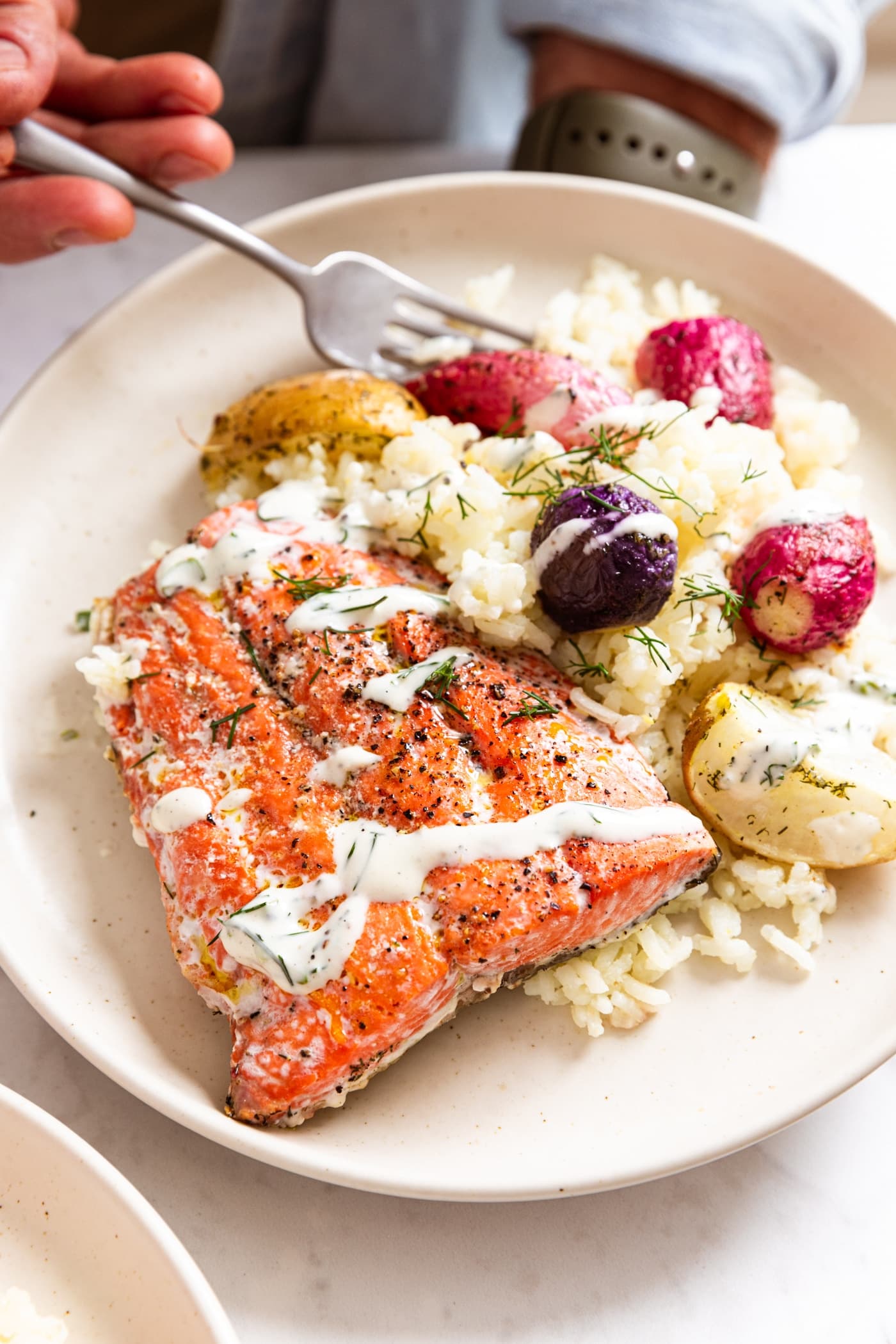 Hand holding white plate filled with salmon with bed of rice topped with sliced radishes and drizzled with homemade buttermilk ranch with fork about to take a piece.