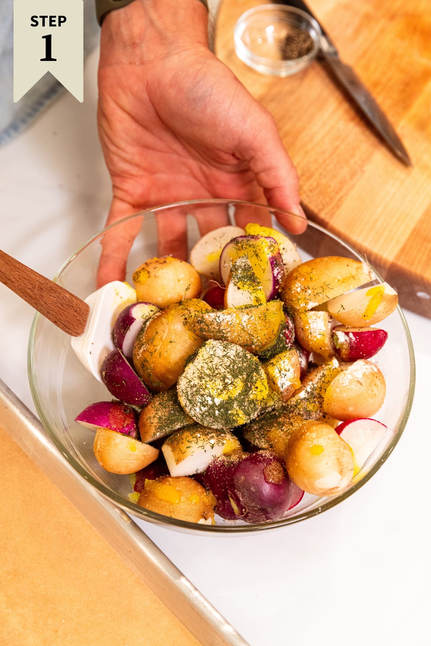 Hand holding glass bowl on white surface filled with sliced radishes sprinkled with dried herbs mixing together with parchment-lined baking sheet to the side.