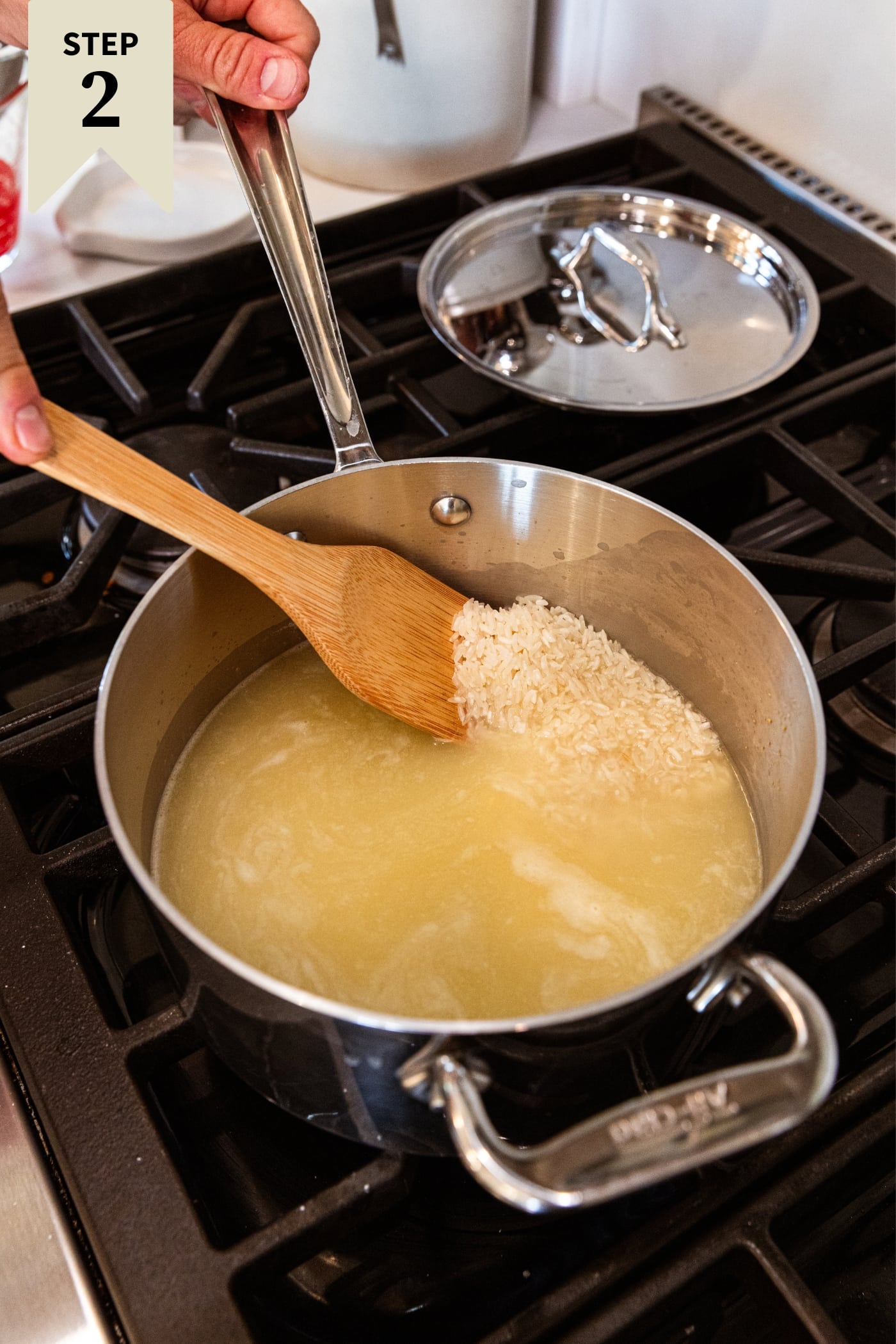 Small kettle sitting on stovetop with hand holding wooden spoon showing uncooked rice in chicken stock.