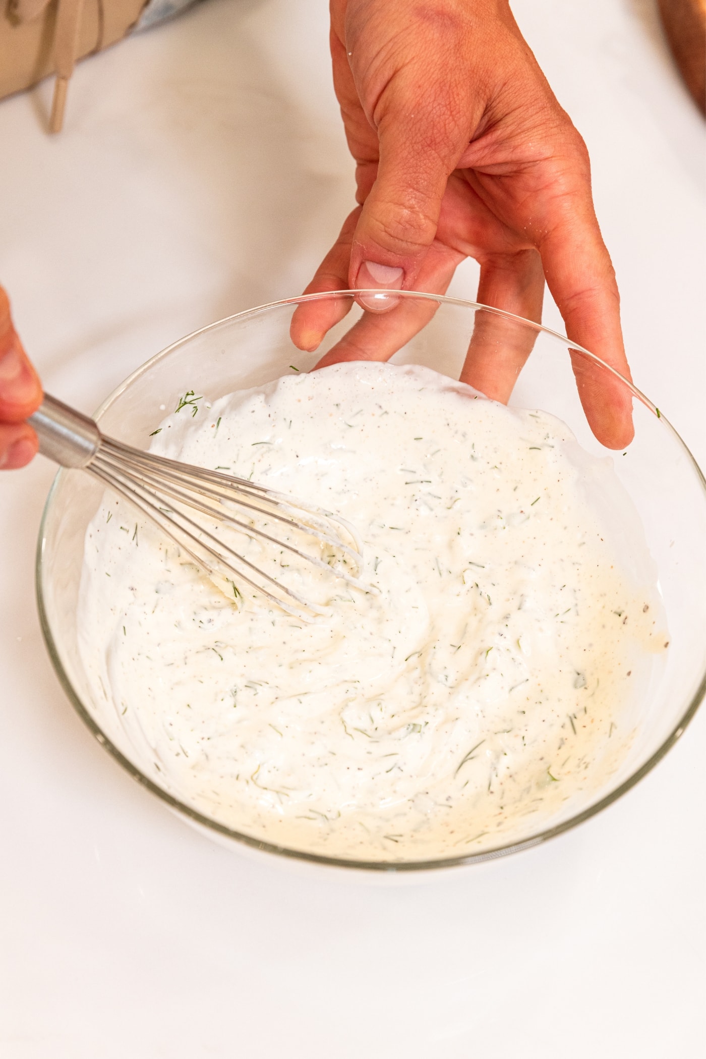 Hand holding the sides of a small glass bowl holding whisk mixing together ranch dressing on white surface.