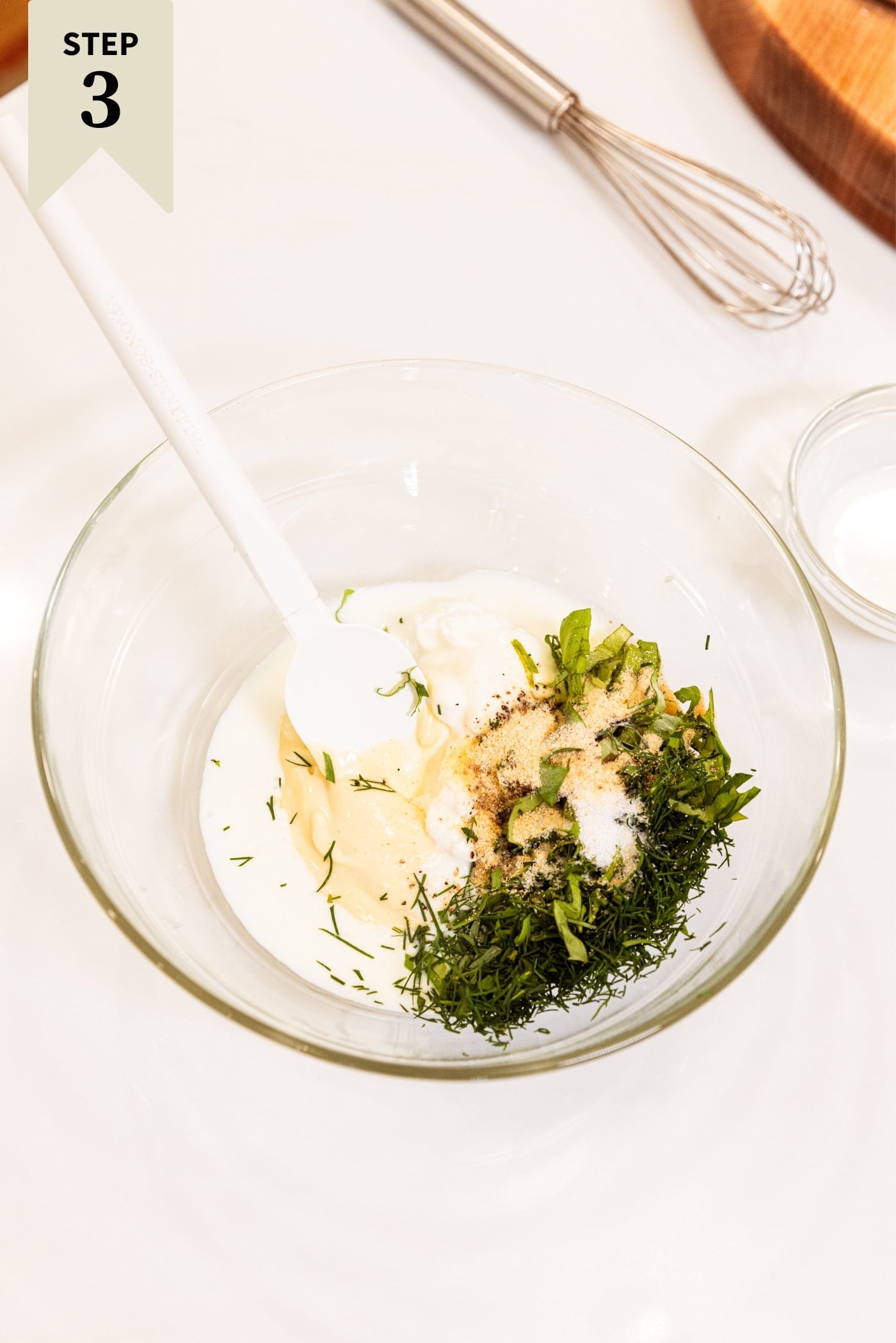 Small glass bowl with buttermilk, minced fresh herbs, Dijon mustard, and more before being mixed together with white spatula all on white surface.