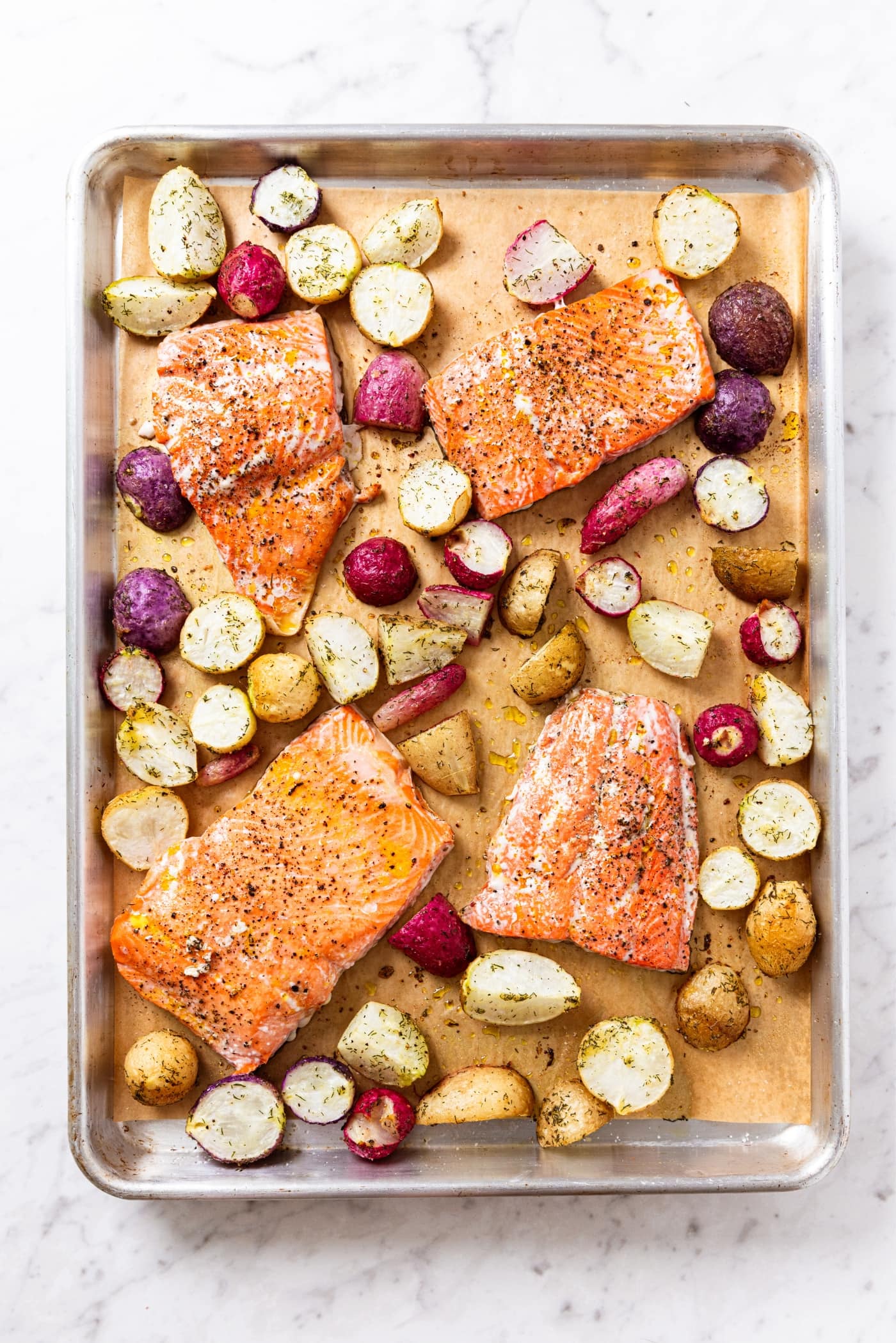 Cooked pieces of salmon resting among sliced cooked radishes on parchment-lined baking sheet all on white marble surface.