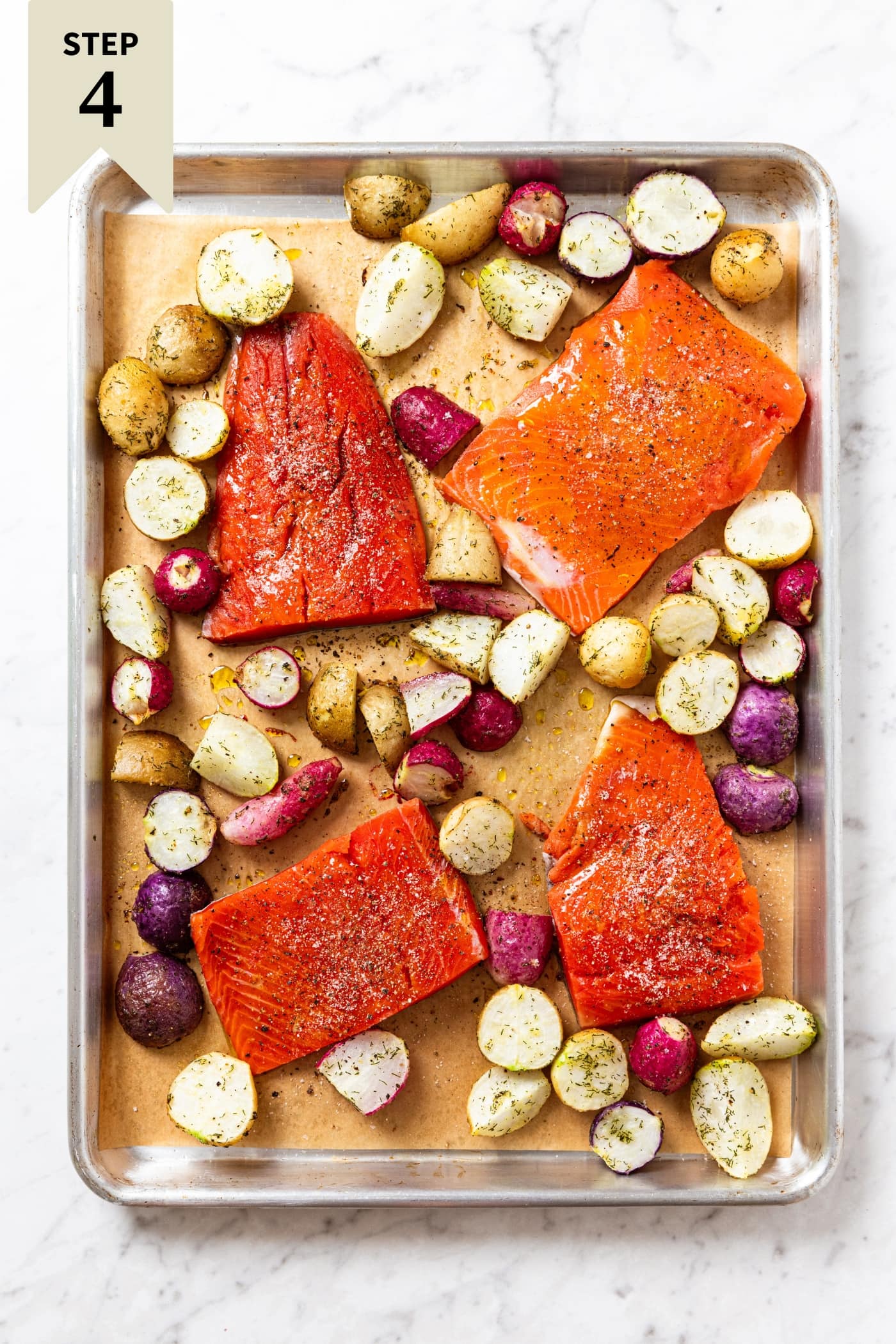Large metal baking sheet sitting on white marble surface with uncooked pieces of salmon resting amongst sliced radishes with dried herbs all over.