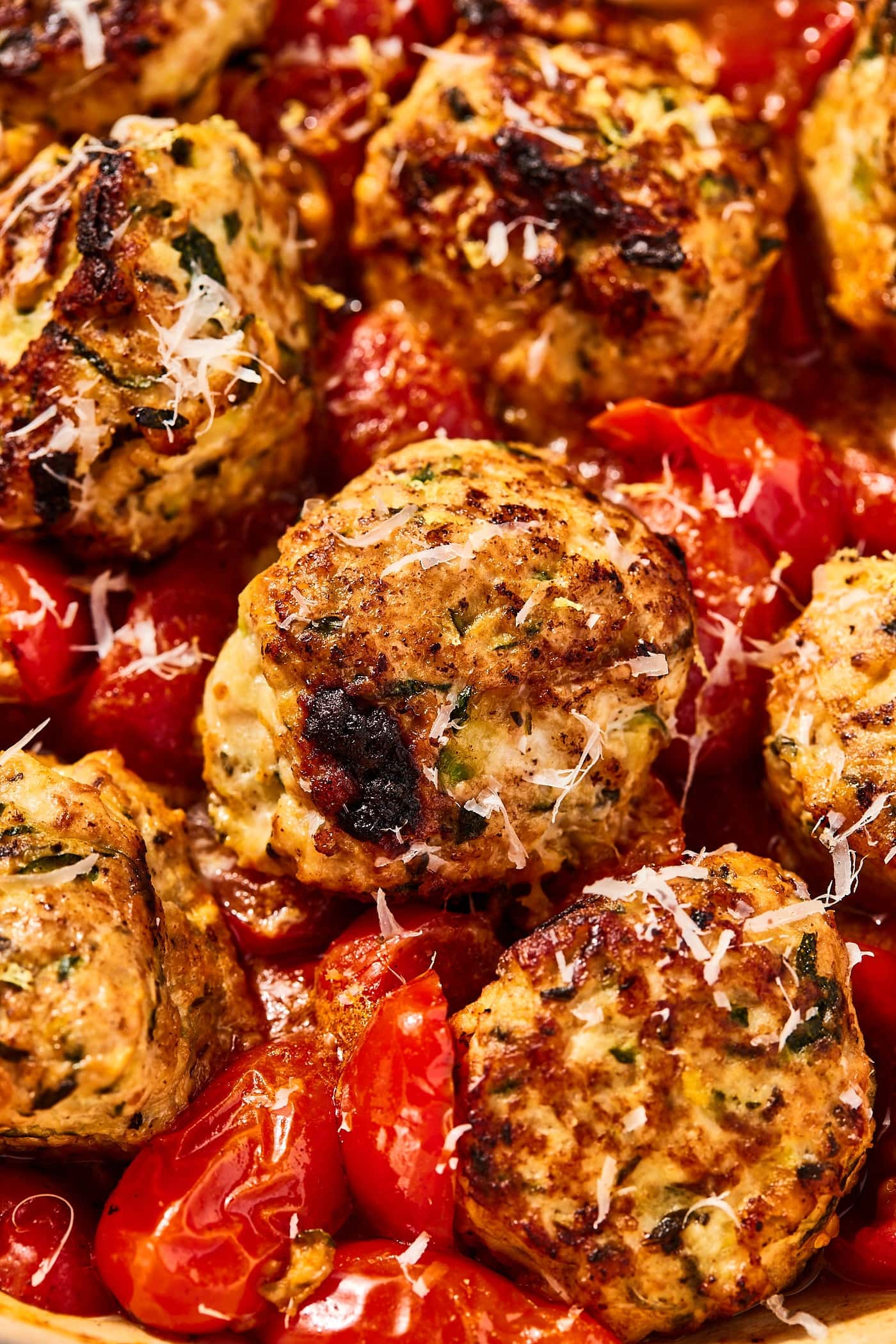 Browned chicken zucchini meatballs sitting in cooked tomatoes with slices of browned garlic around all in skillet.