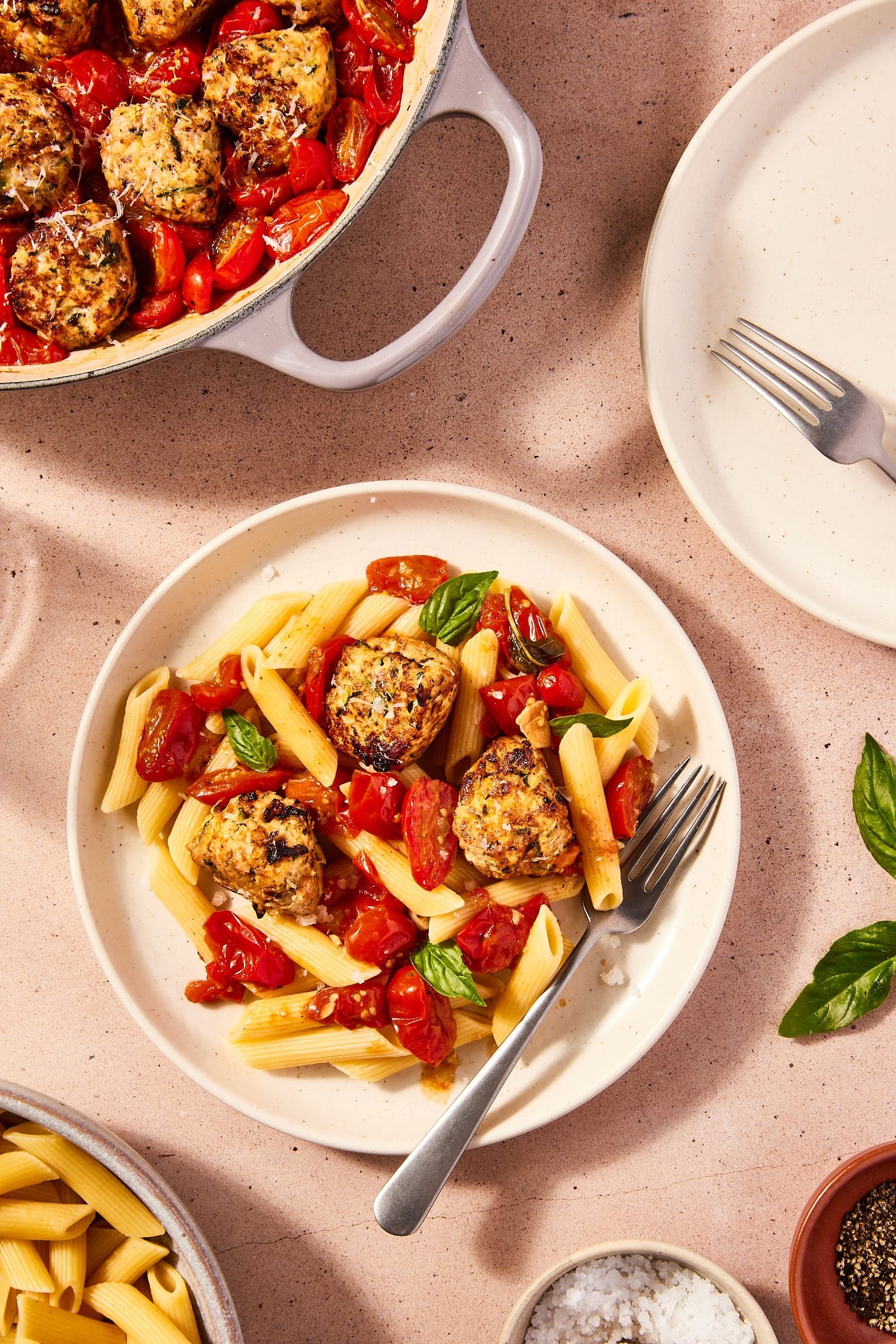 Round white plate with chicken zucchini meatballs and tomatoes nestled on top of penne noodles with fresh basil torn on top and skillet with rest of meatballs to the side.
