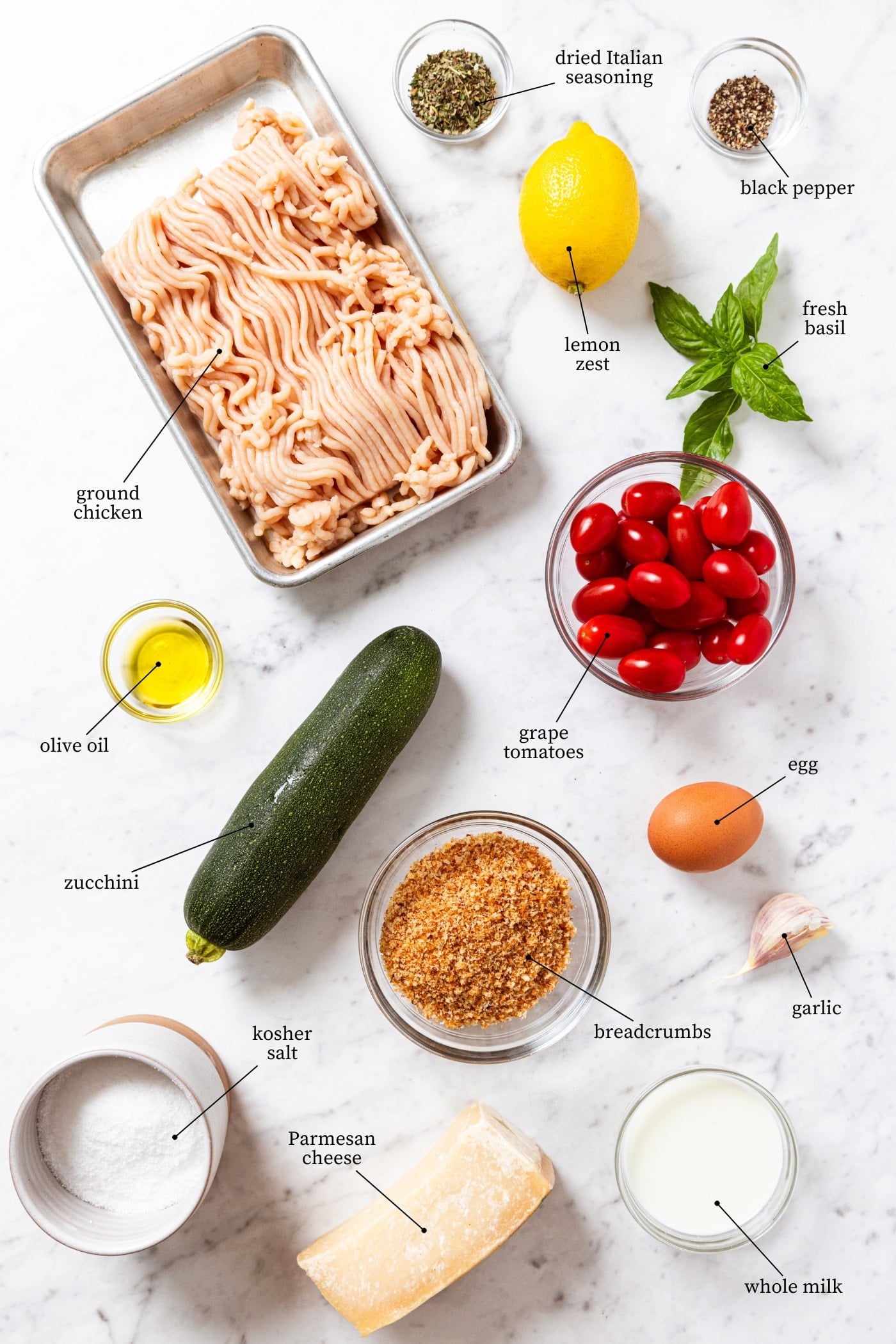 White marble surface filled with all ingredients needed to make chicken zucchini meatballs including raw ground chicken, zucchini, grape tomatoes, basil, lemon, bread crumbs, spices, and more.