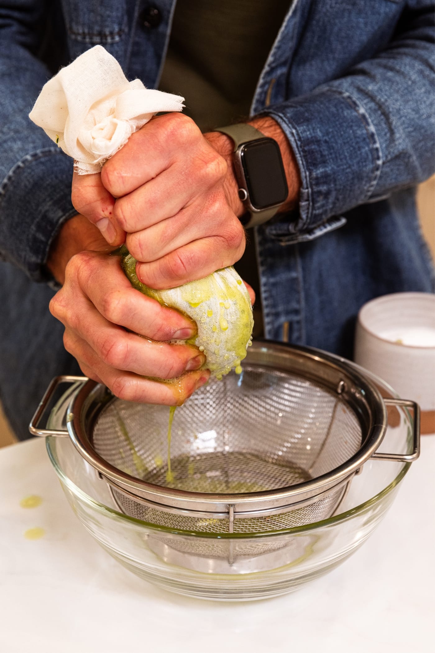Two hands squeezing cheesecloth filled with shredded zucchini over strainer with bowl underneath it.