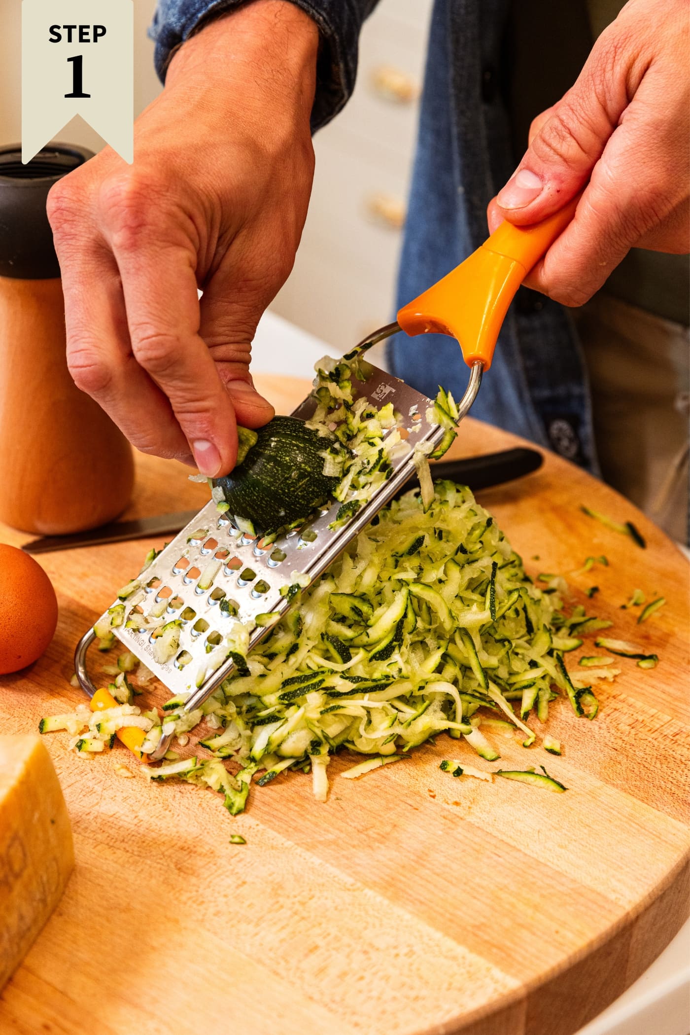 Hand using grater to grate zucchini into a pile on a wood cutting board with pieces of cheese to the side.