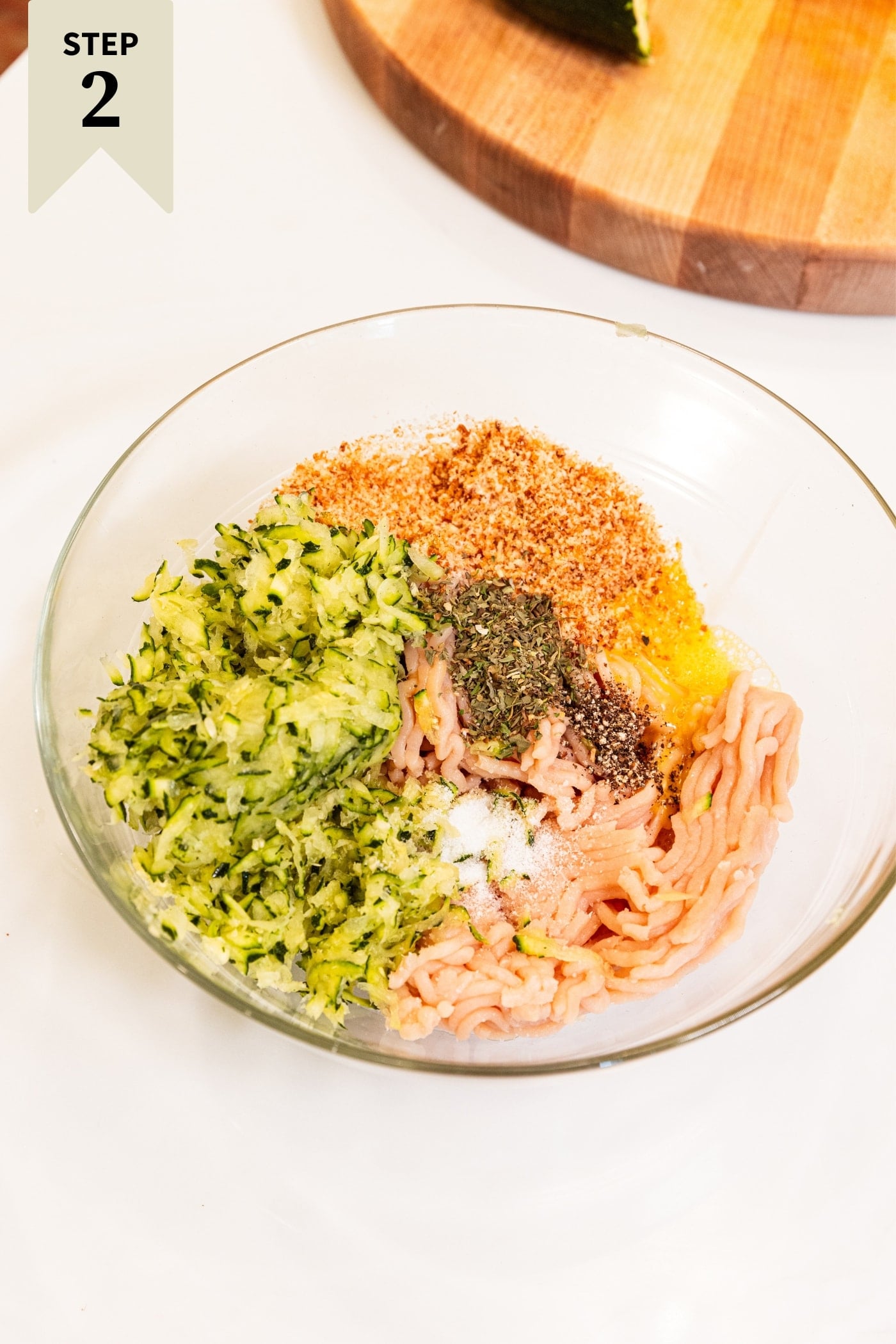 Glass bowl filled with all ingredients needed for chicken zucchini meatballs before being mixed all on white countertop.