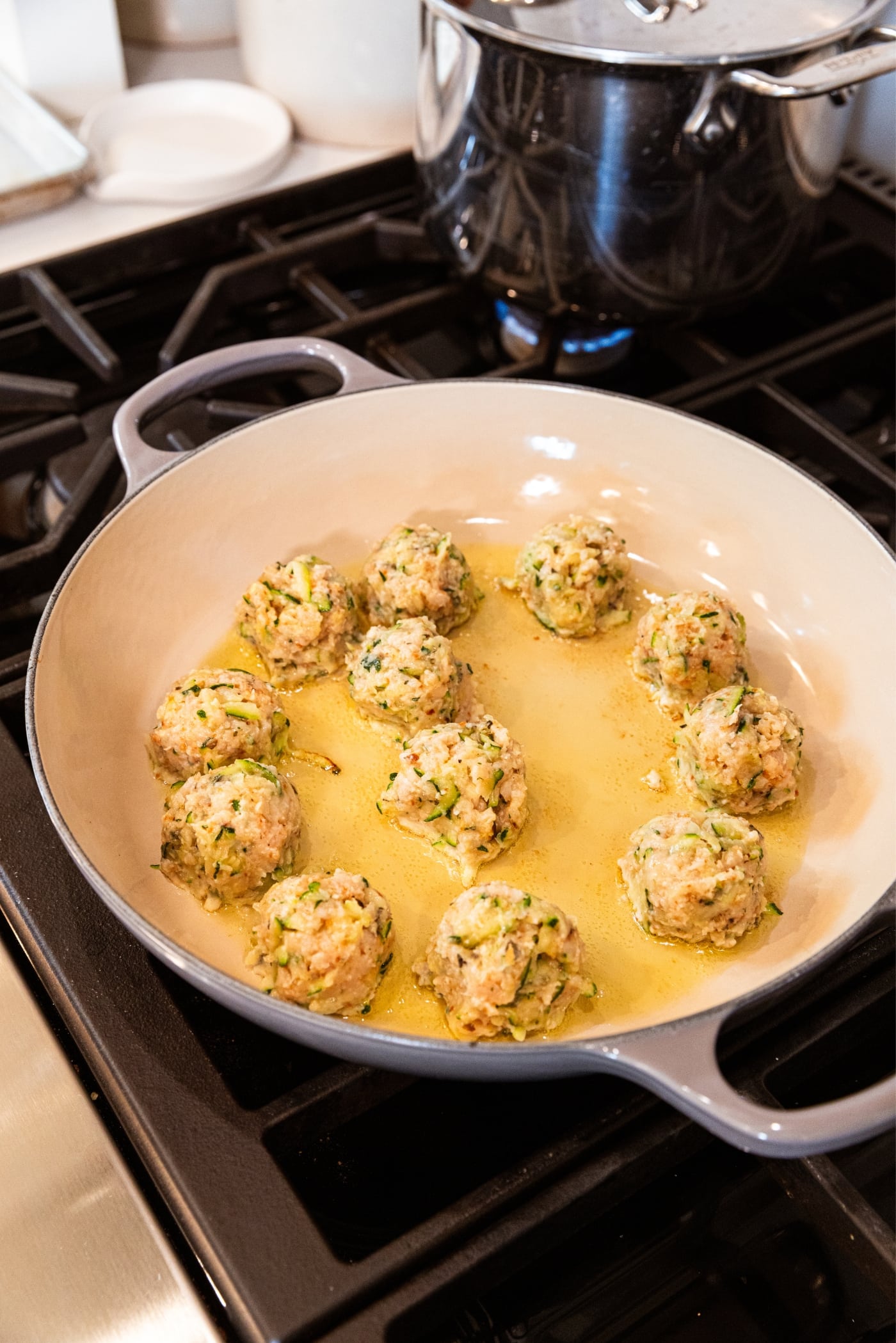 Round chicken zucchini meatballs sitting in oil in a gray skillet on the stovetop.