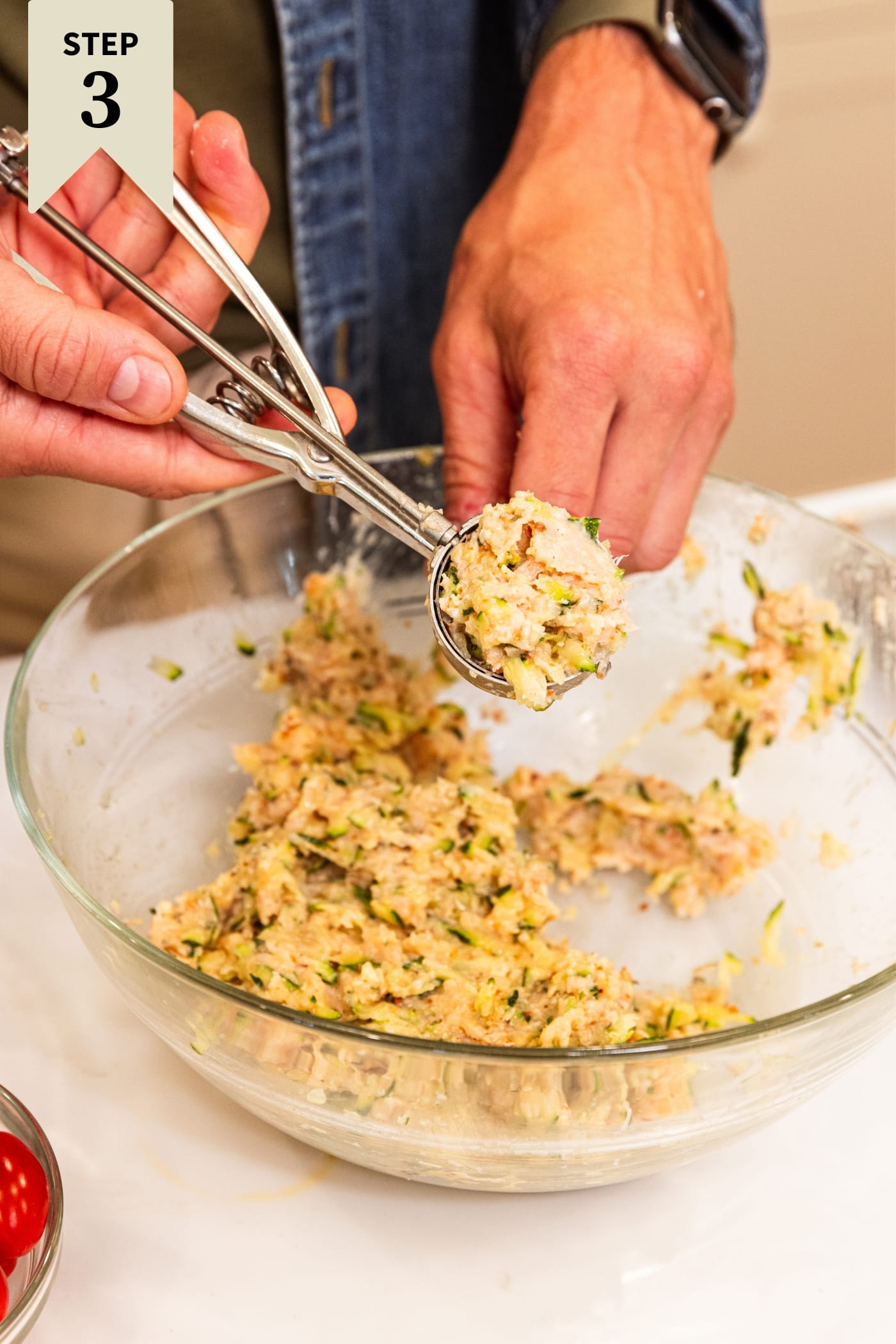 Hand using scoop to scoop up ground chicken mixture from glass bowl sitting on white countertop.