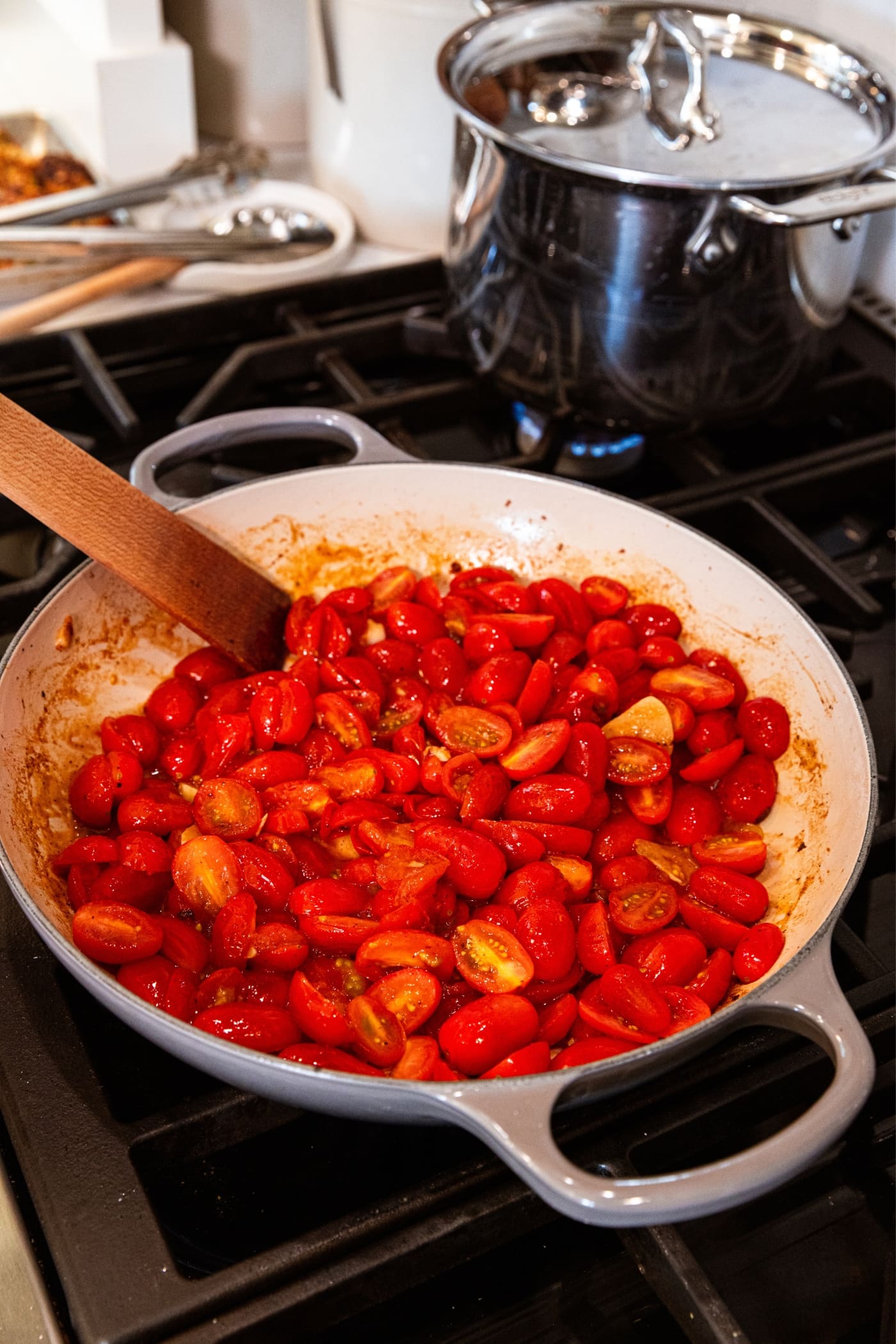 Gray skillet on stovetop with wooden spatula stirring sliced grape tomatoes with their juices escaping.