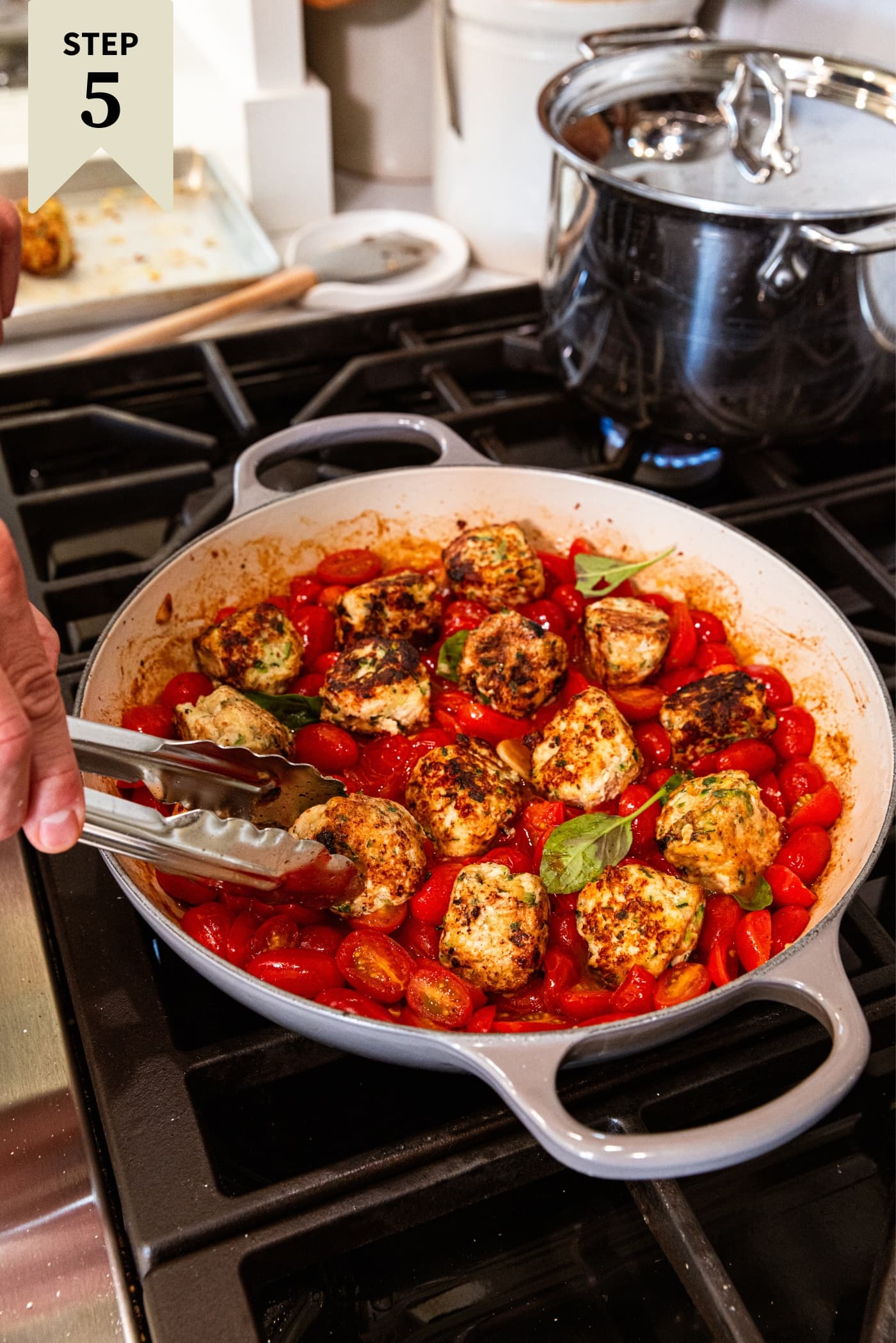 Hand holding tongs placing cooked and browned chicken zucchini meatballs in tomato sauce with fresh basil leaves in skillet on stovetop.