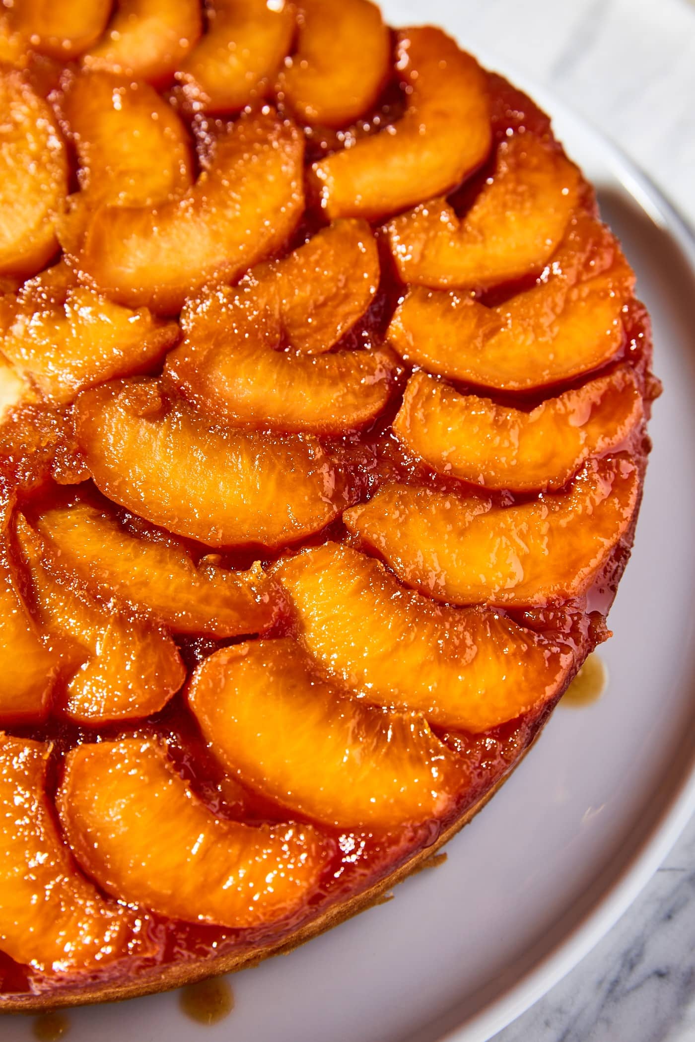 Upside down peach cake sitting on white serving platter with slices of peaches mixed with caramel sauce on top.