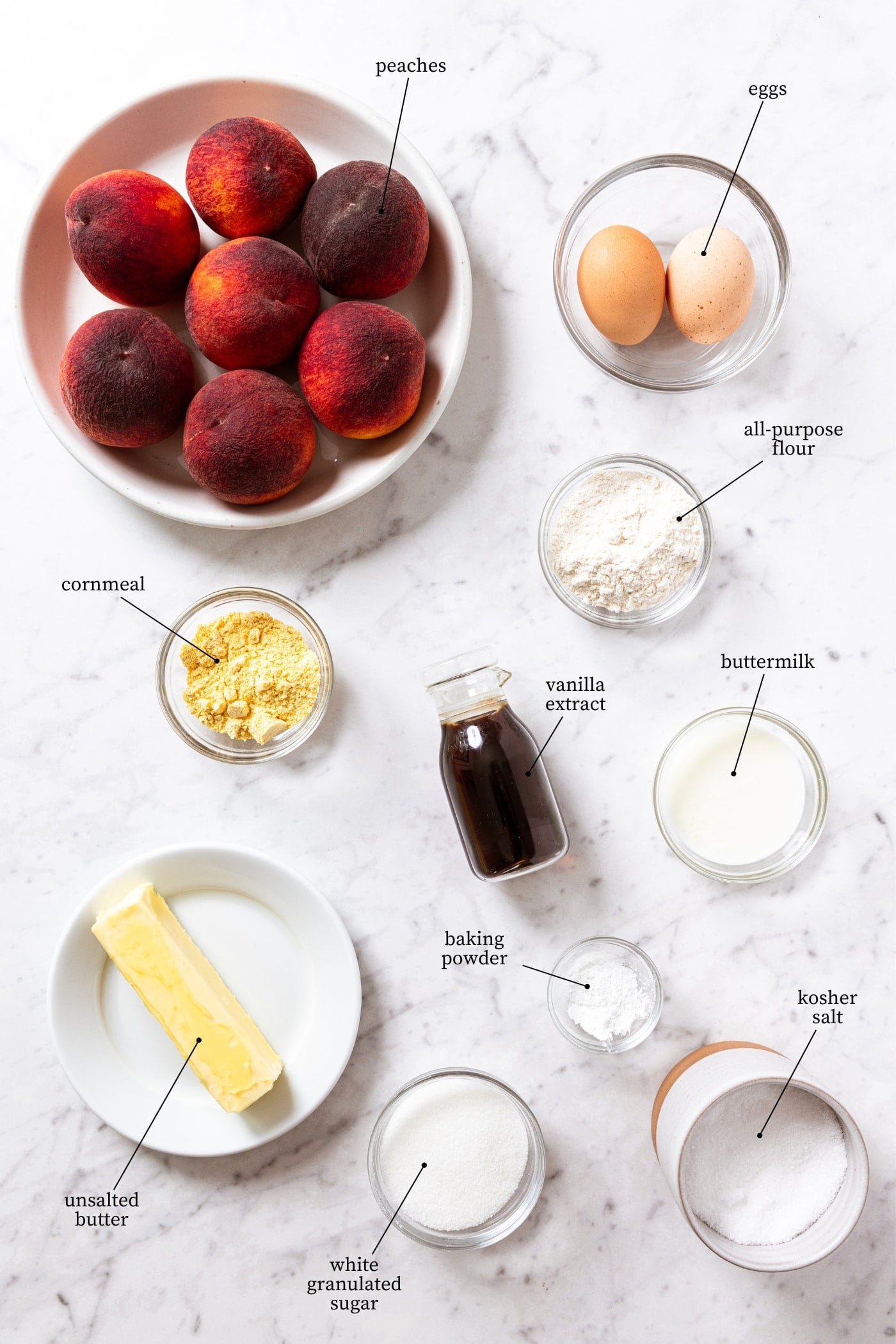 Whit marble surface filled with ingredients needed to make an upside down peach cake including peaches, butter, eggs, sugar, flour, vanilla extract, and more.