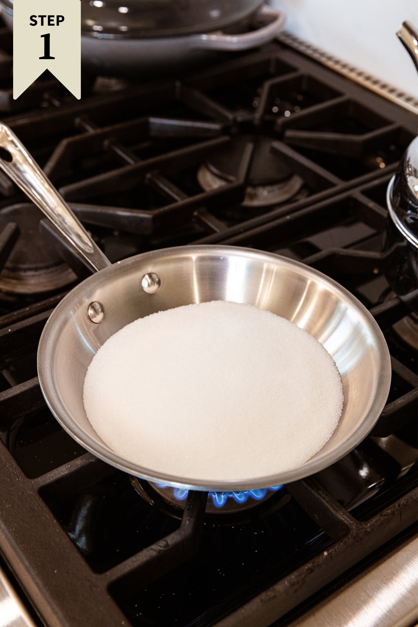 Metal skillet sitting on stovetop with raw white sugar before melted over the heat.