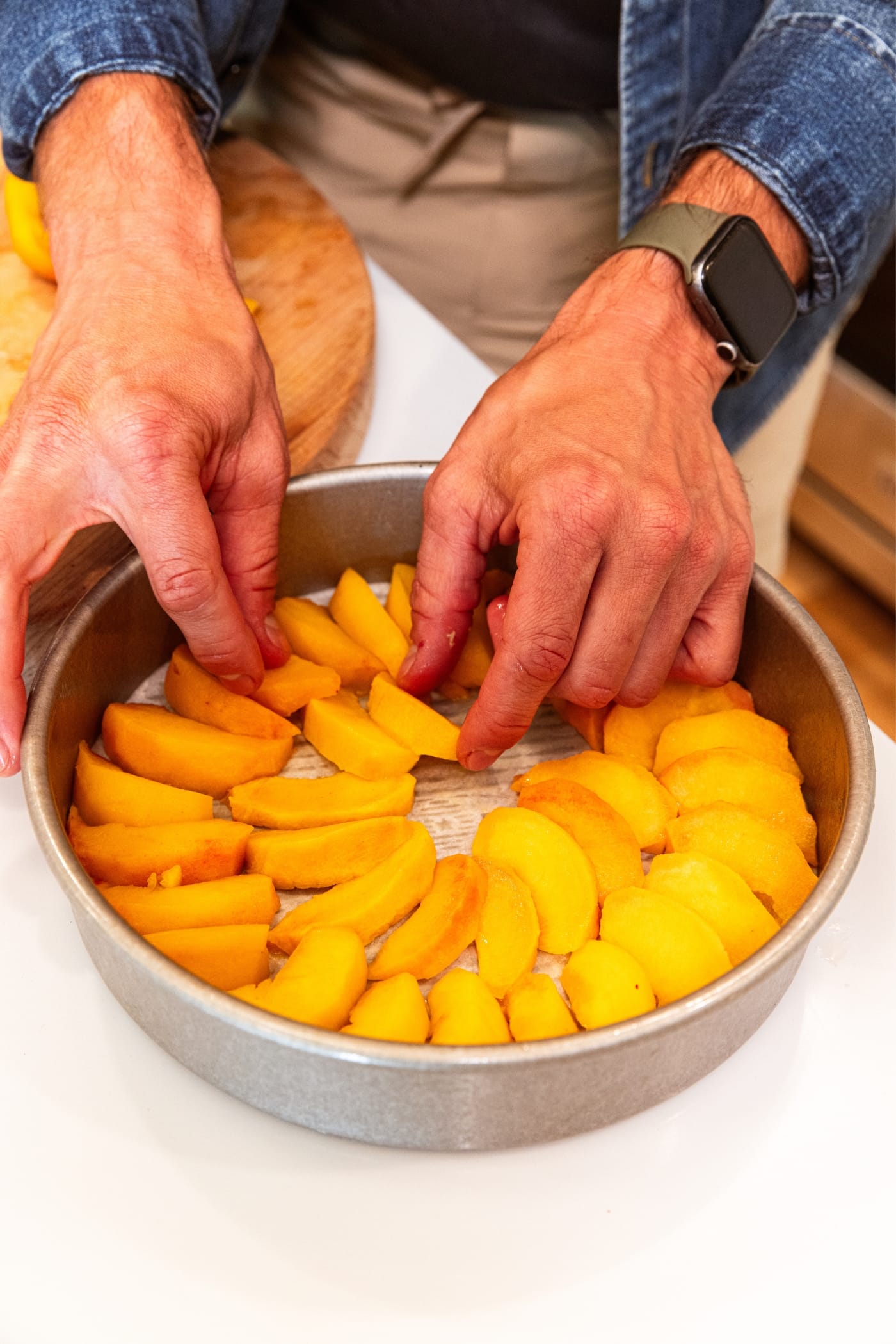 Two hands placing wedges of peaches into a round metal baking pan with parchment round along the bottom all on white surface.