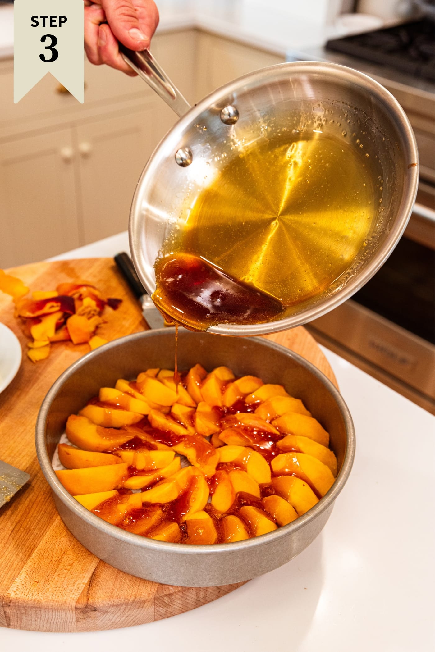 Hand holding skillet with melted caramel sauce pouring over the top of sliced peaches sitting in metal circular pan on cutting board.