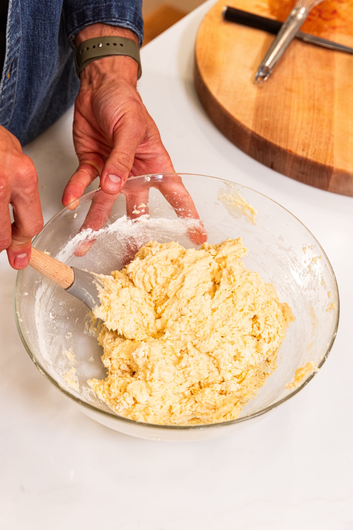 Hand using plastic spatula to fold together batter ingredients for a cake all on white surface.