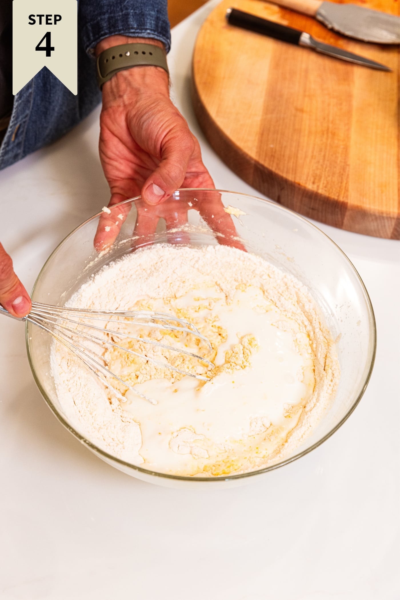 Hand holding whisk mixing together dry ingredients in large glass metal bowl sitting on white surface.