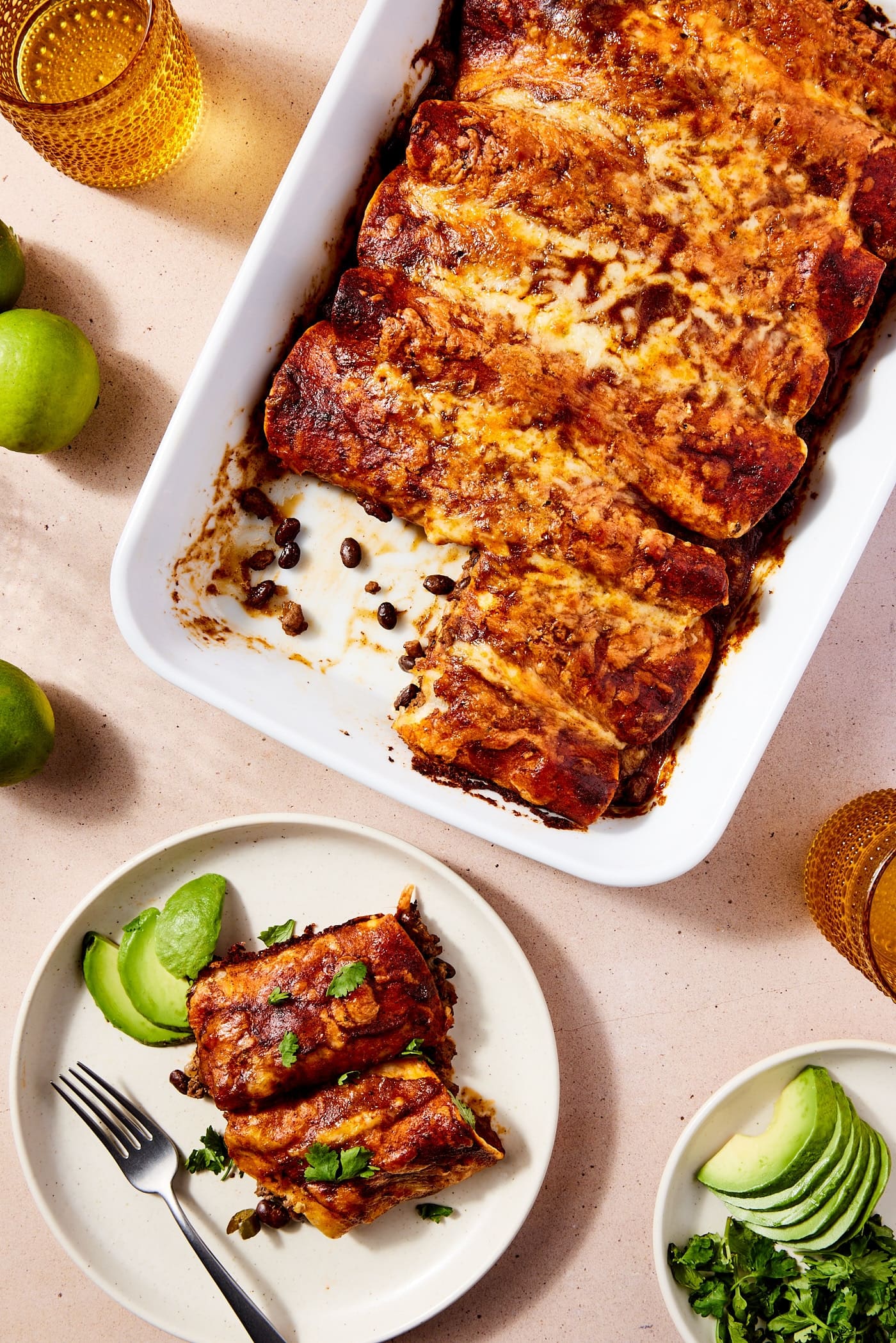White baking dish filled with baked beef enchiladas with a plate with two pieces of tortilla with fork resting on the plate as well along with sides such as cilantro and avocado.