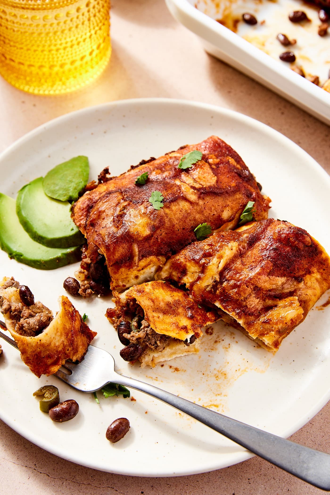 White plate with two flour tortilla beef enchiladas with fork slicing a serving from one of the tortillas with slices of avocado in the background.