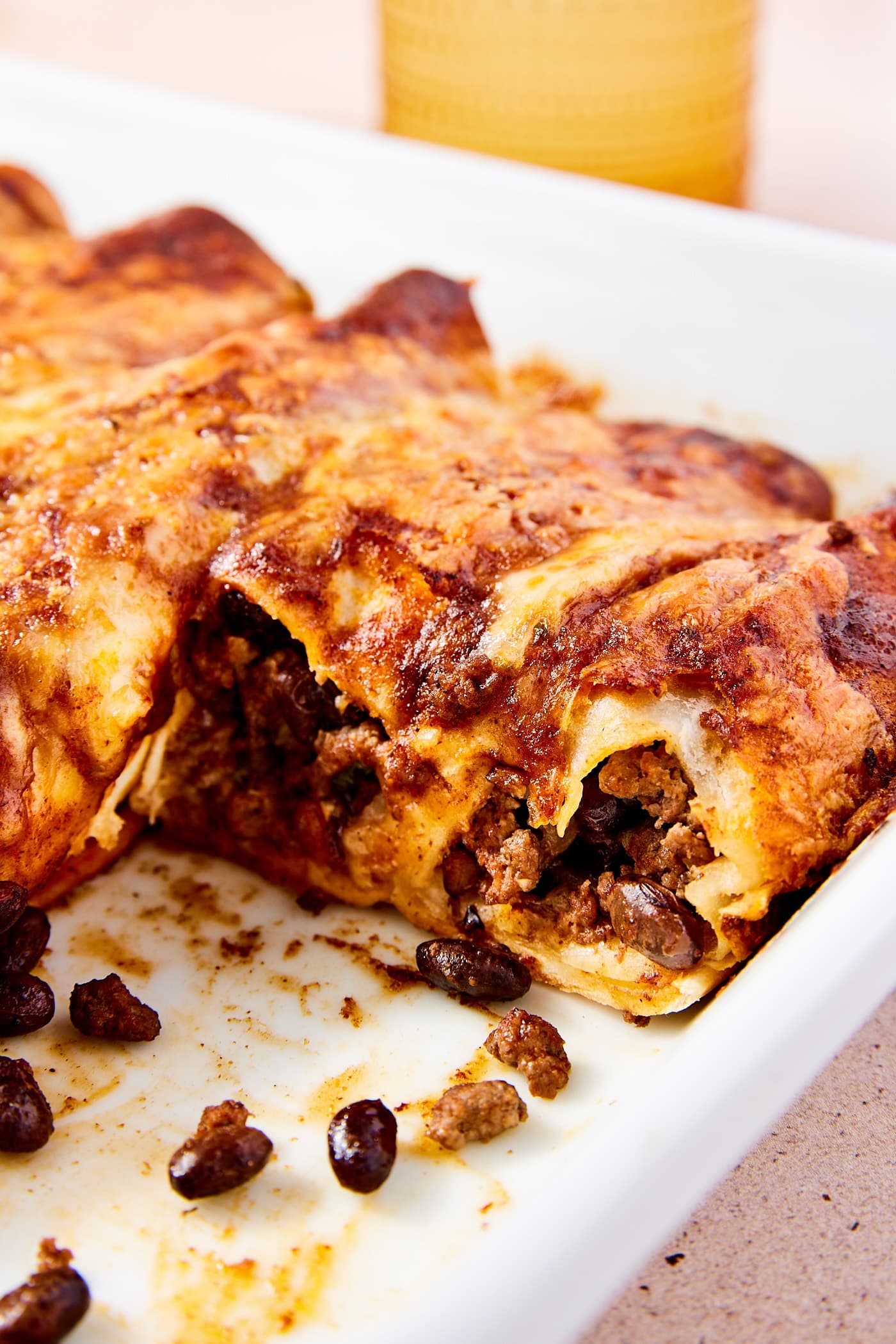 White rectangular baking dish filled with beef enchiladas with two tortilla pieces missing showing the interior texture of the enchiladas with beans spilling out.
