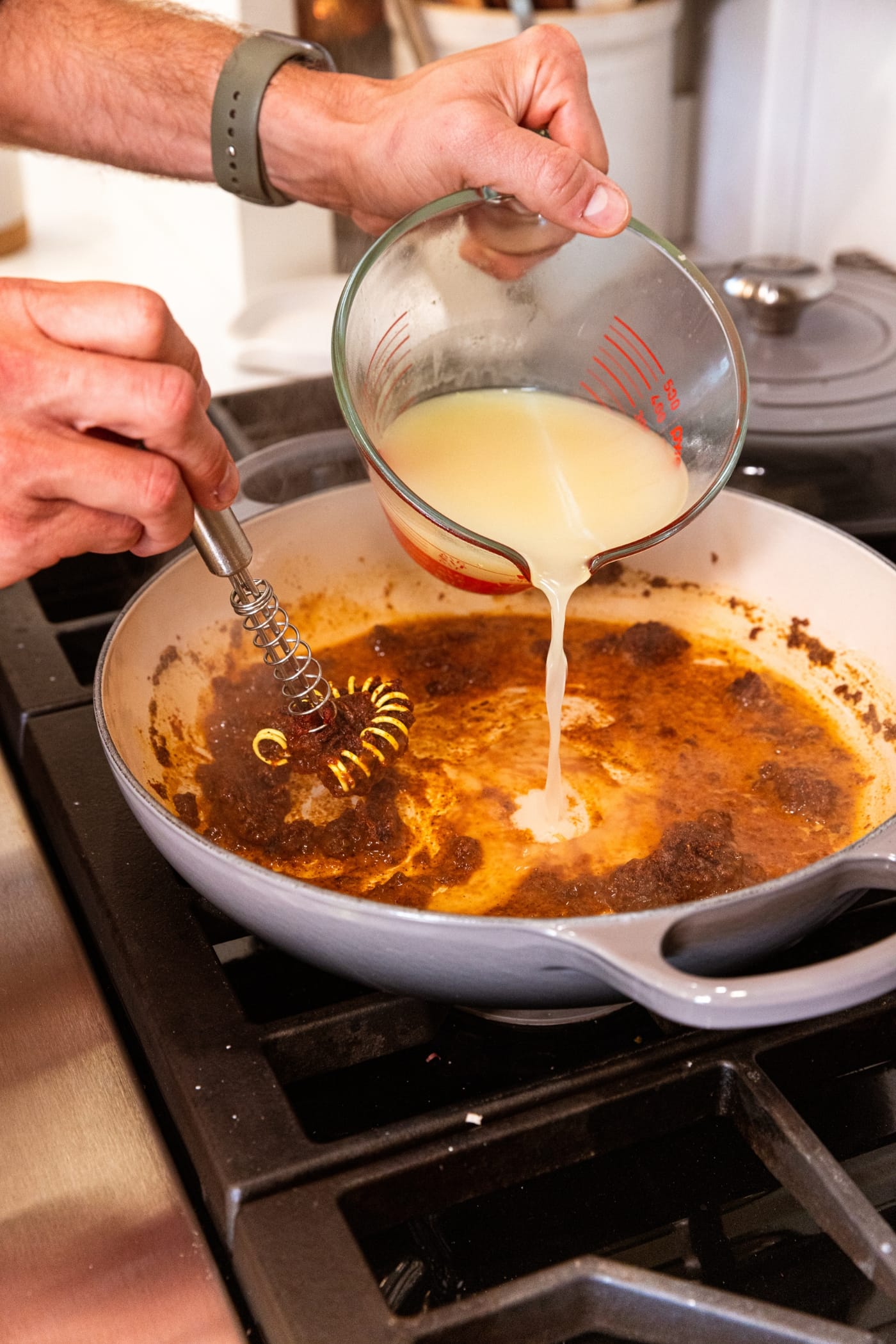 Hand holding glass measuring cup pouring chicken stock into simmering mixture of spices and flour in a pan on the stove.