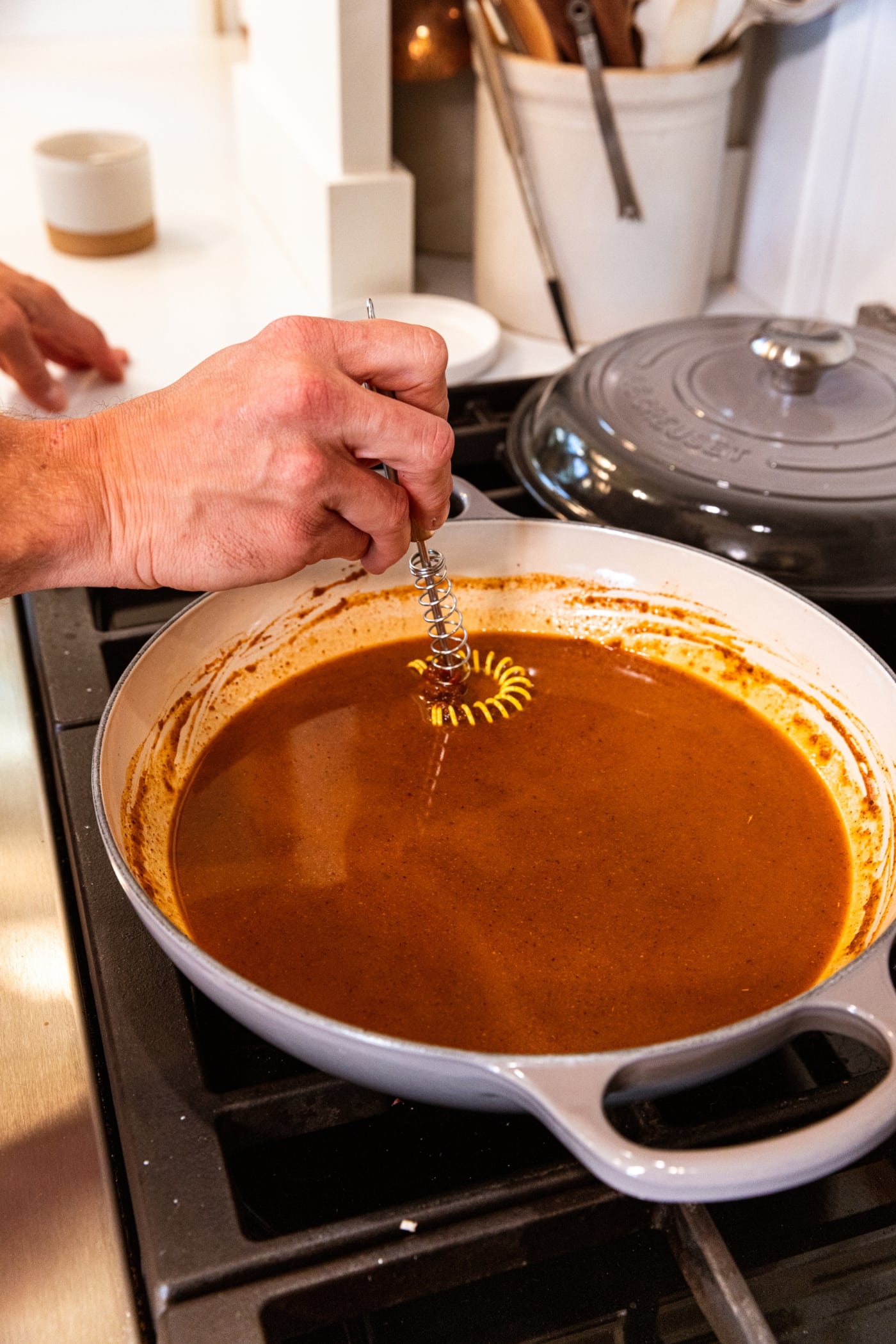Hand holding small whisk mixing together enchilada sauce in a pan on the stovetop.