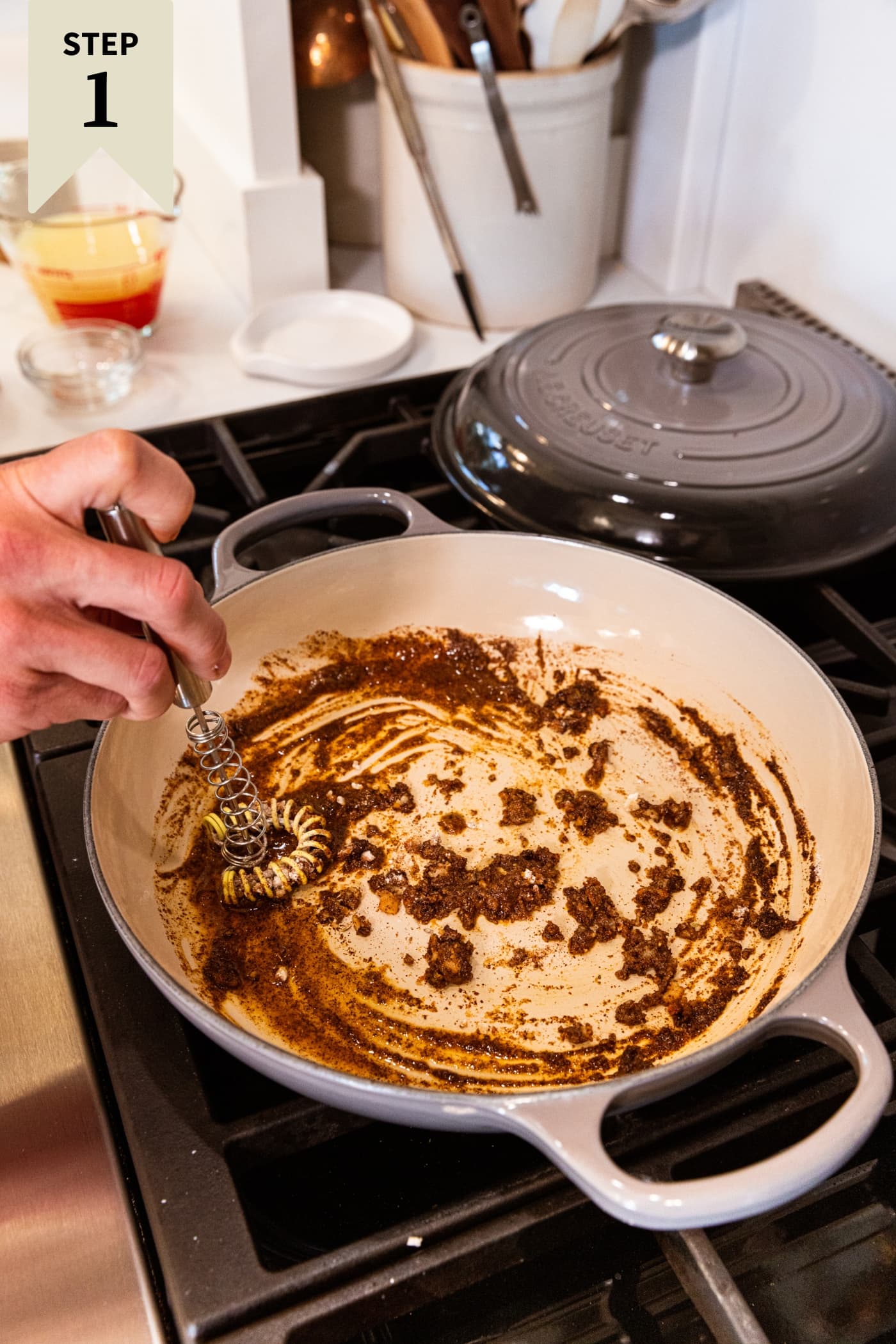 Hand holding small flat whisk mixing together tomato paste with spices and flour in a pan on the stove.