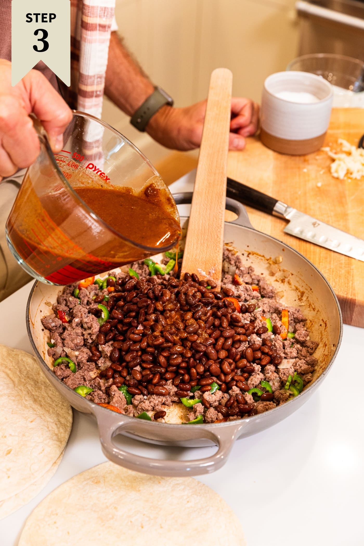 Hand holding glass measuring cup mixing in enchilada sauce on top of cooked browned meat with black beans on top all in glass bowl.
