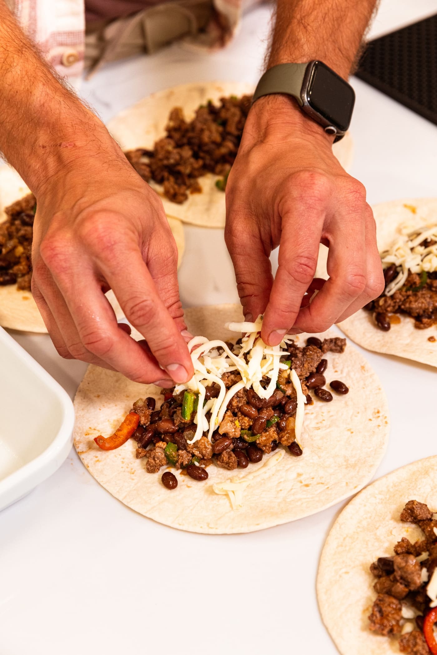 Two hands placing shredded cheese on top of meat mixture sitting on a flour tortilla on a white surface.
