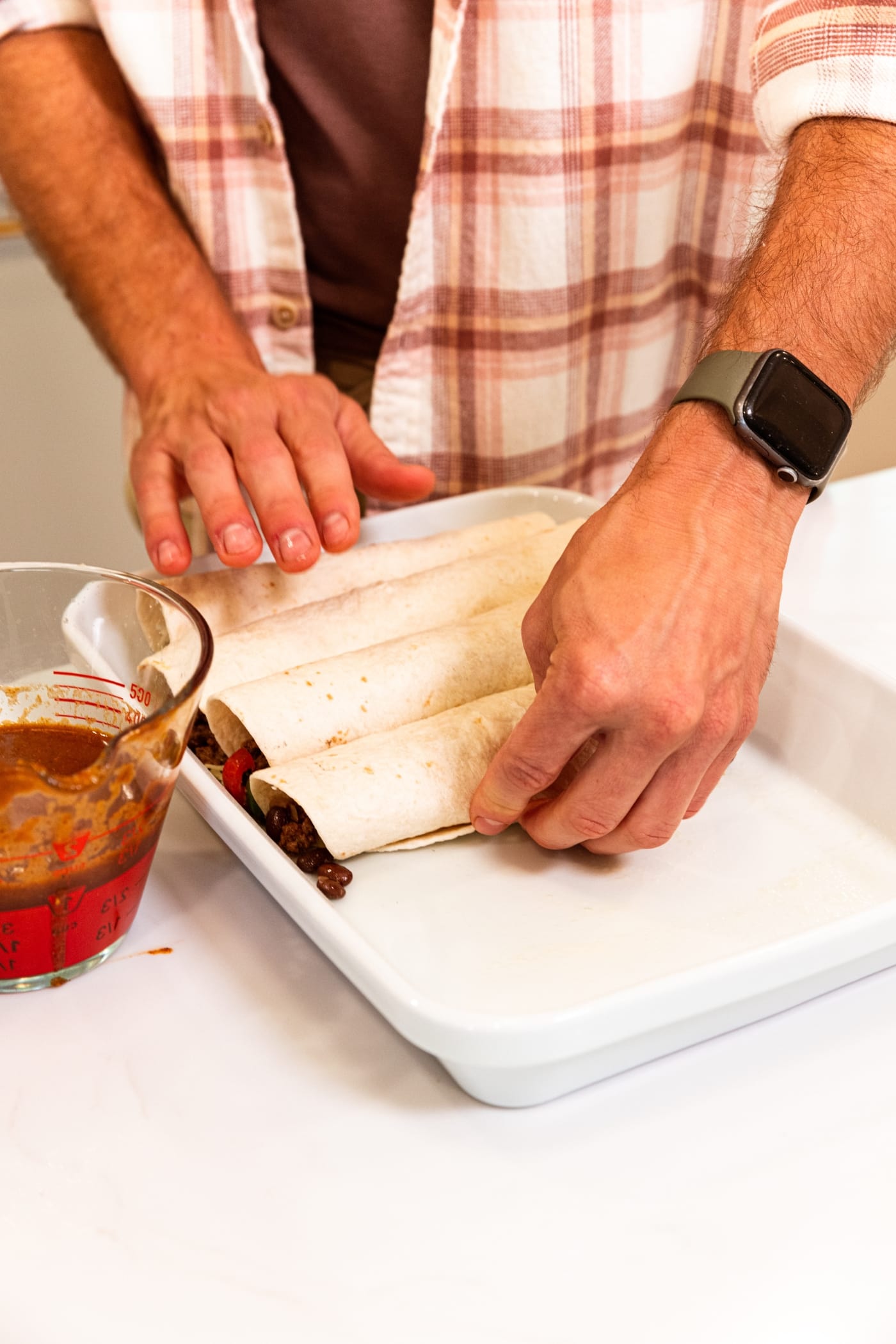 Hands placing a rolled flour tortilla filled with meat mixture alongside other tortillas in a white baking dish.