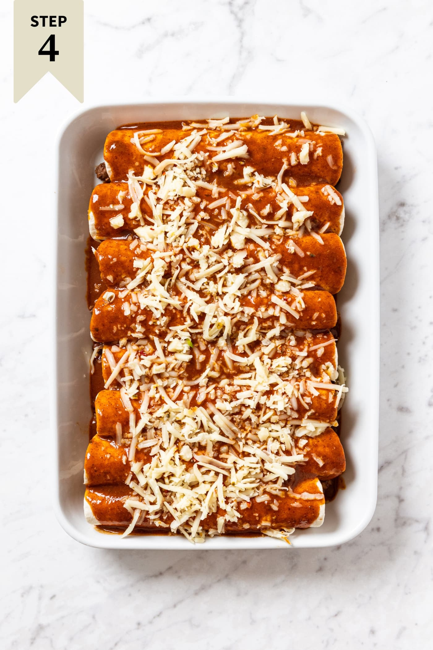 Unbaked beef enchiladas with shredded cheese on top sitting in white baking dish before going into the oven.