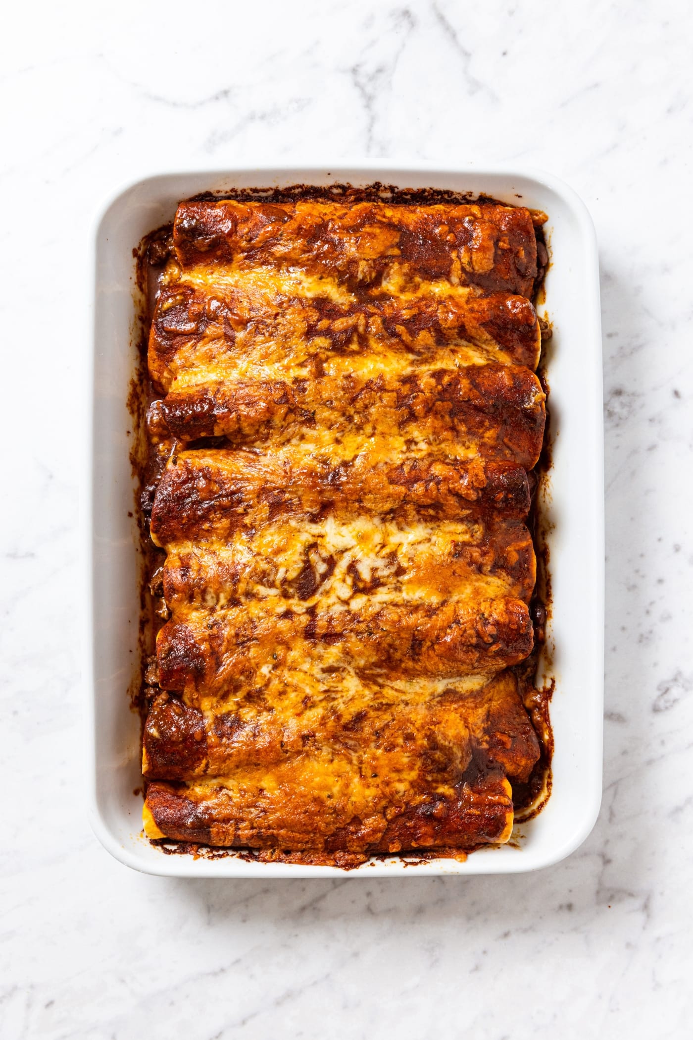 Baked enchiladas cooling in a white baking dish sitting on white marble surface.