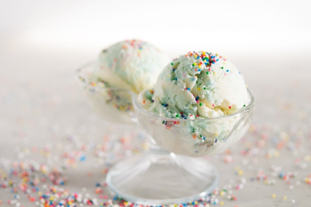 Two scoops of blue and white ice cream with colorful sprinkles in a glass container in front of a white background.