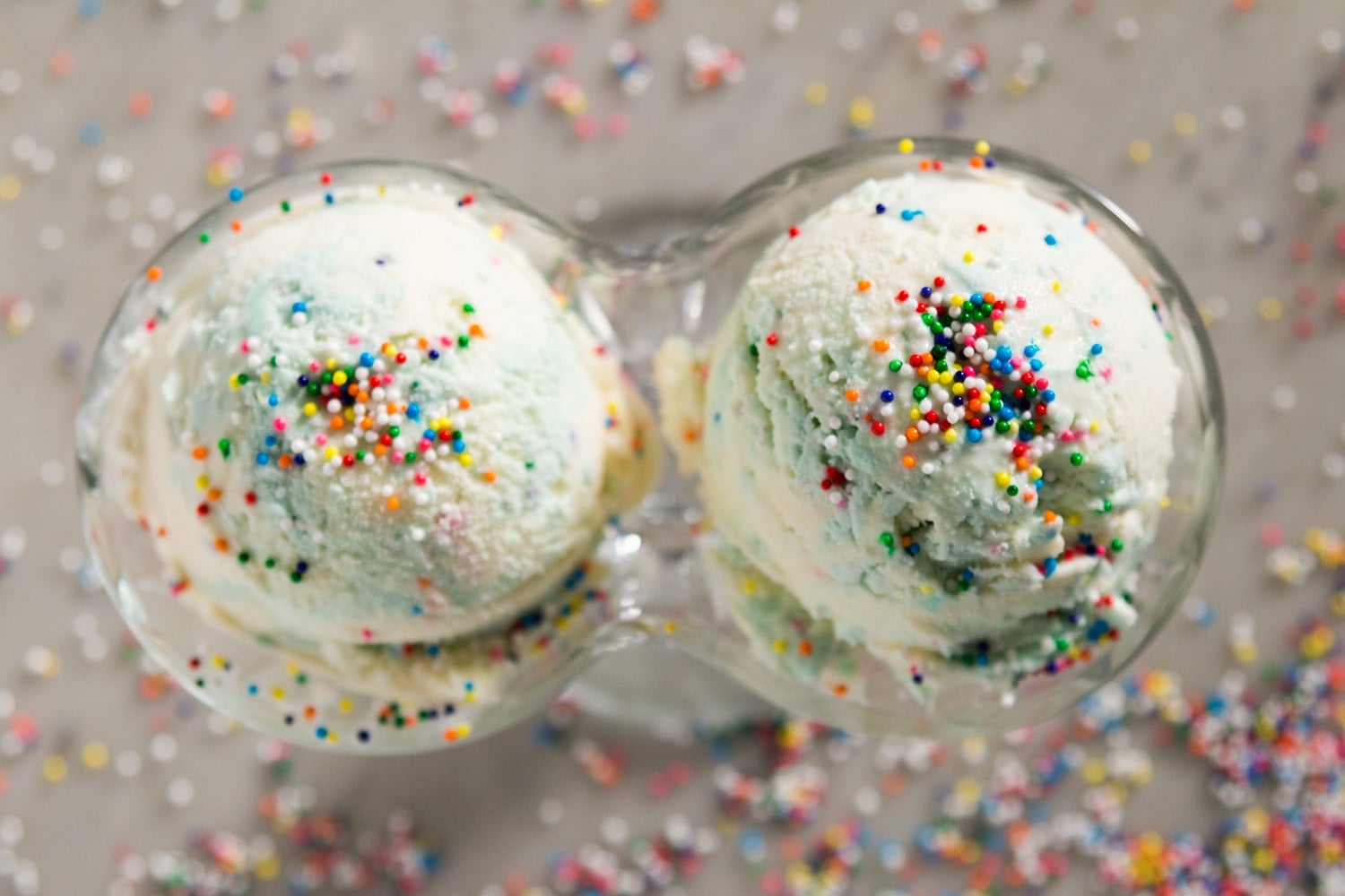 A top-down view of two scoops of blue and white ice cream with sprinkles in a glass container on a gray surface. 