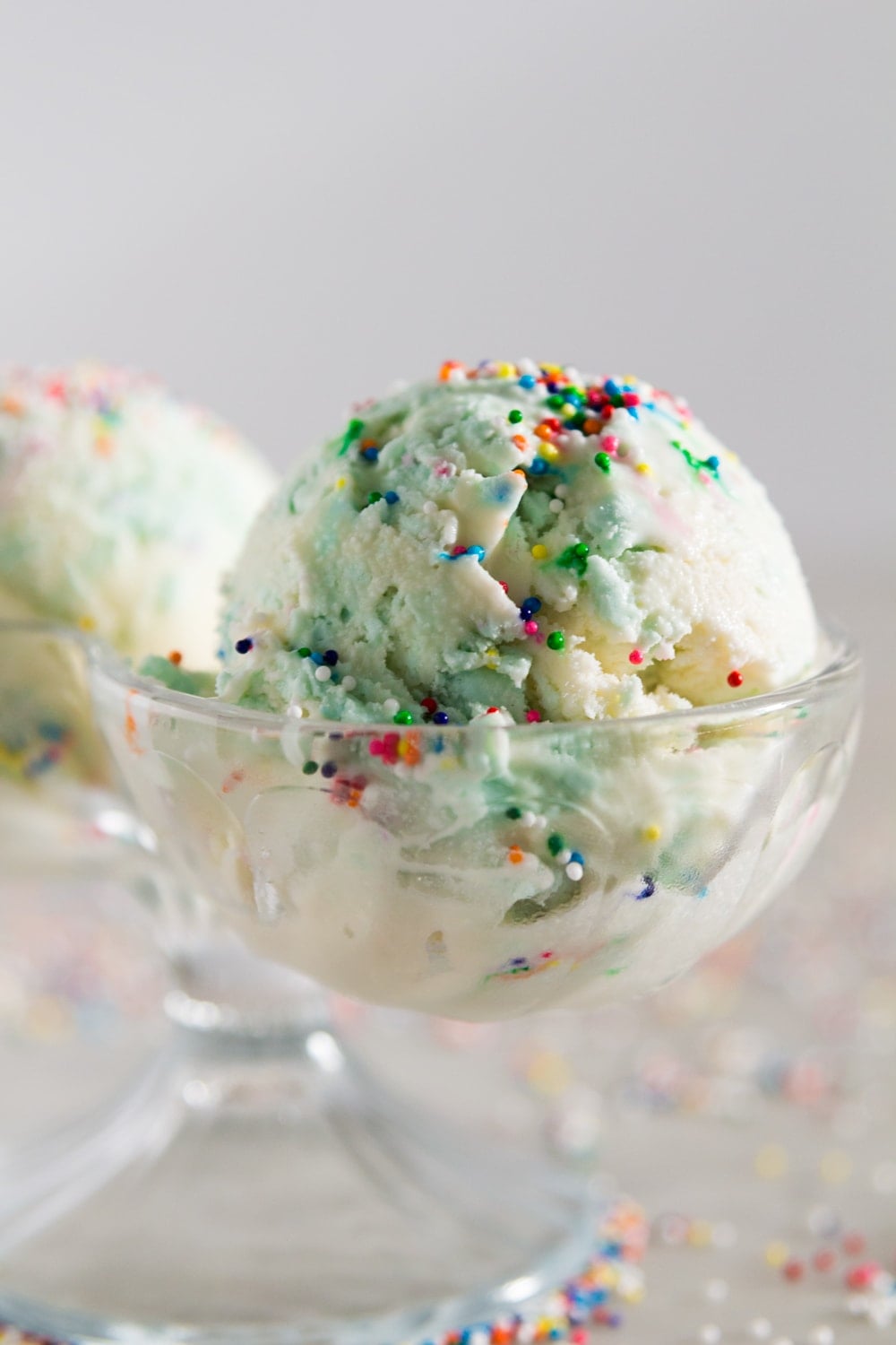 A glass container holding a blue and white scoop of ice cream with sprinkles on top in front of a white background. 