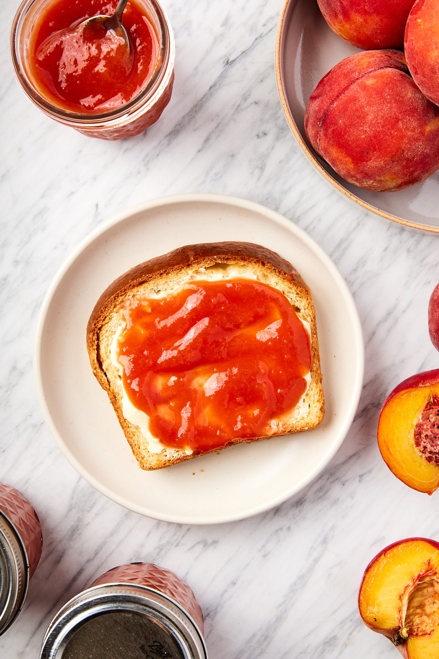 White marble surface with plate with slice of bread topped with butter and peach butter with oranges all around along with more jars of butter.