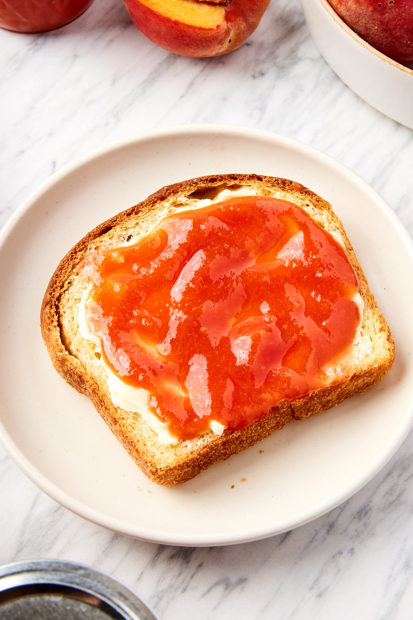 White marble surface with white plate with slice of bread slathered with butter and peach butter.