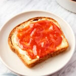 White marble surface with piece of bread spread with butter and peach butter all on white plate with peaches all around.